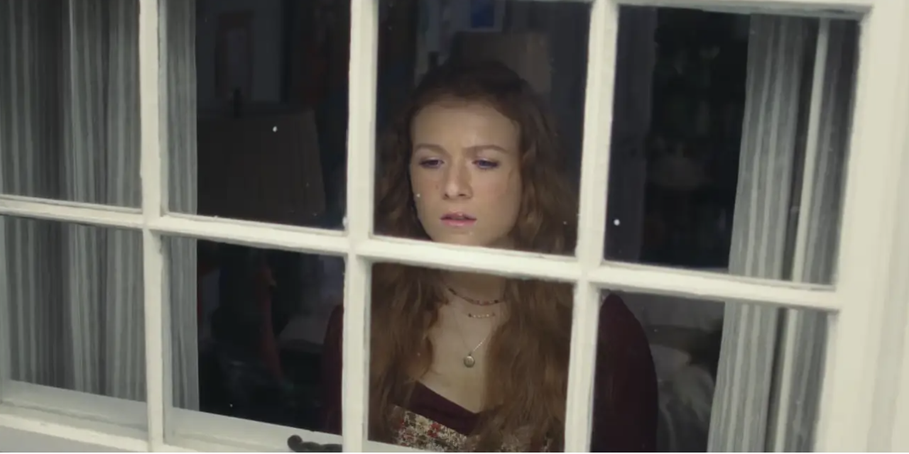 A young Lily looks pensively out of a multi-paned window from inside a room, appearing contemplative