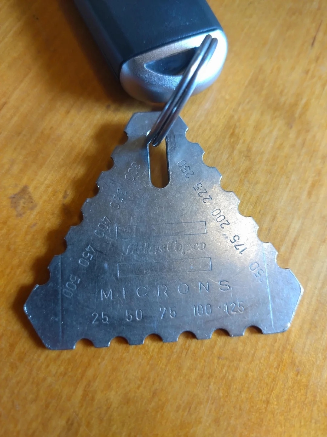 Triangular metal gauge tool labeled "Microns" with various measurements hanging from a keyring attached to a dark handle on a wooden surface