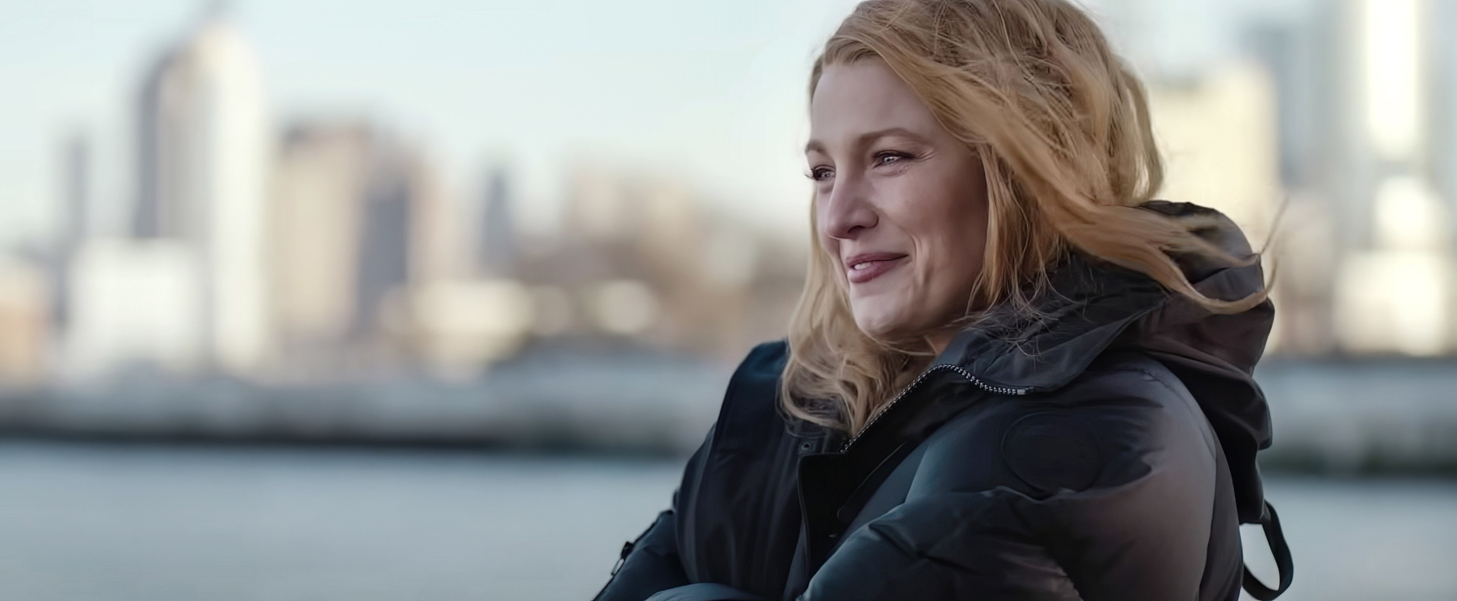 A smiling woman with wavy hair and a black winter coat is outside with a city skyline in the background