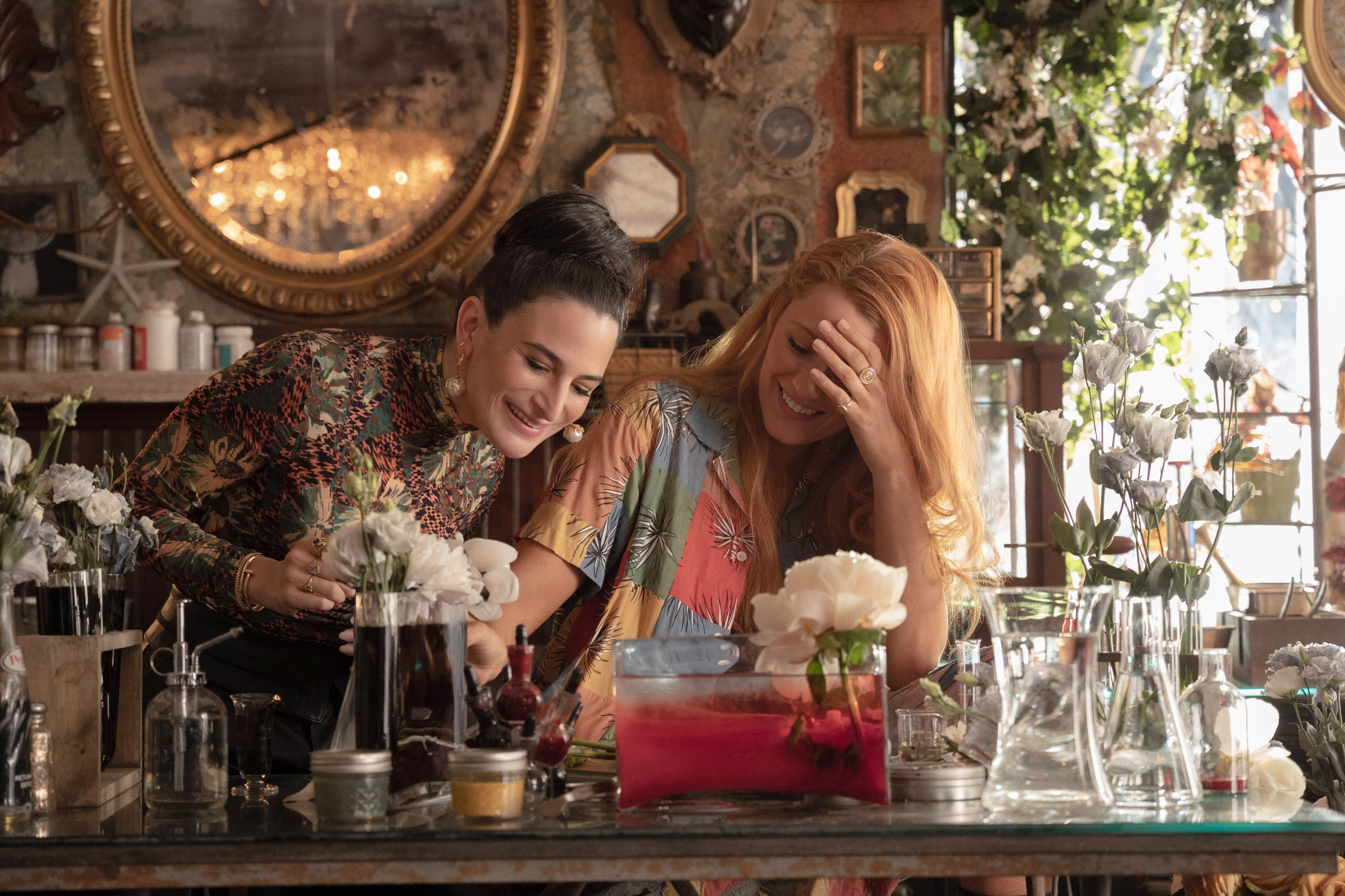 Jenny Slate and Blake Lively laugh together at a table filled with flowers and laboratory glassware in a vintage-styled room