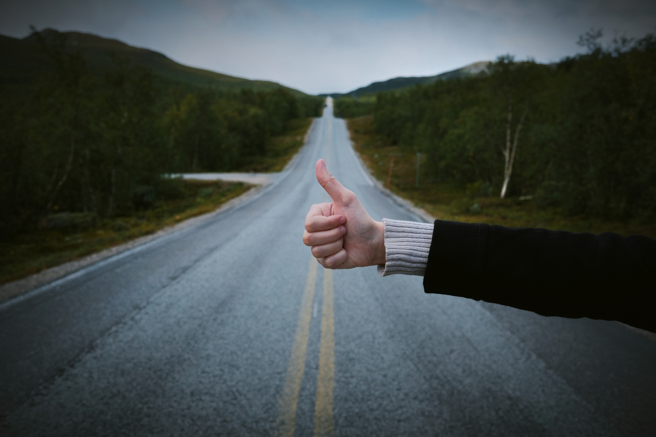 A person's hand is giving a thumbs-up gesture while standing on an empty, straight road surrounded by trees and mountains
