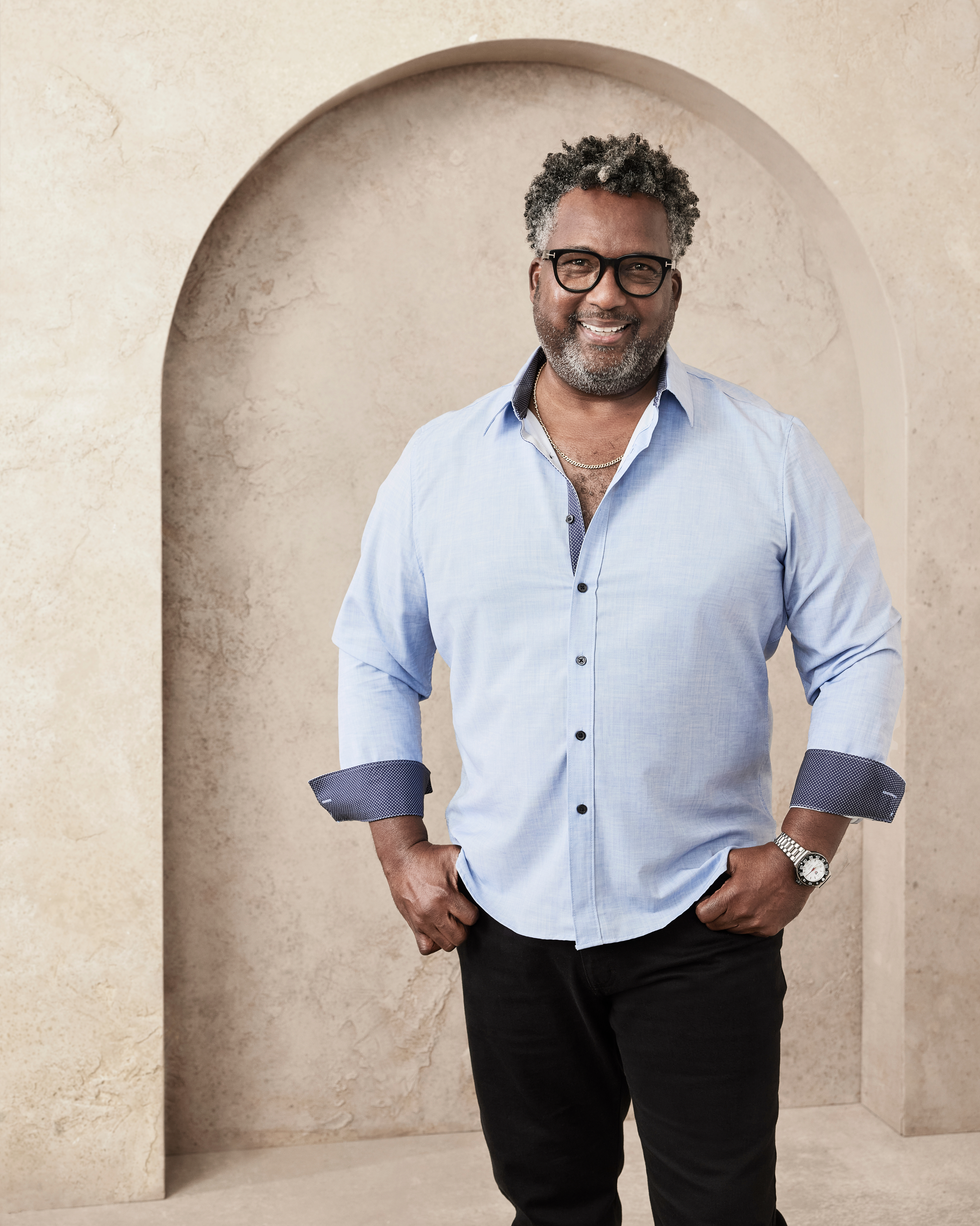 A man with glasses, wearing a light shirt with dark cuffs and black pants, smiles in front of a beige textured background