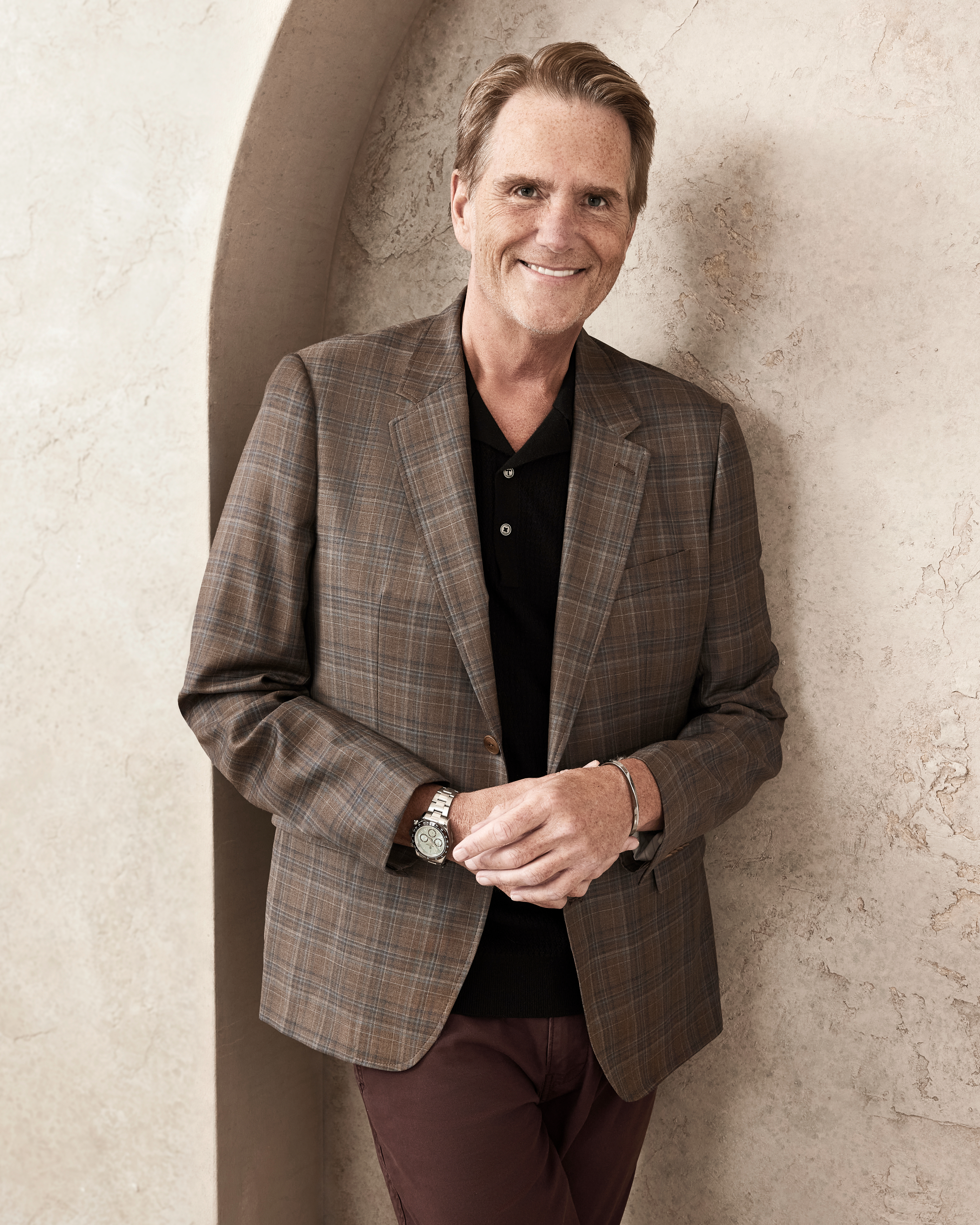 Rob Loughran smiles, leaning against a wall, wearing a plaid blazer over a dark shirt with dark trousers