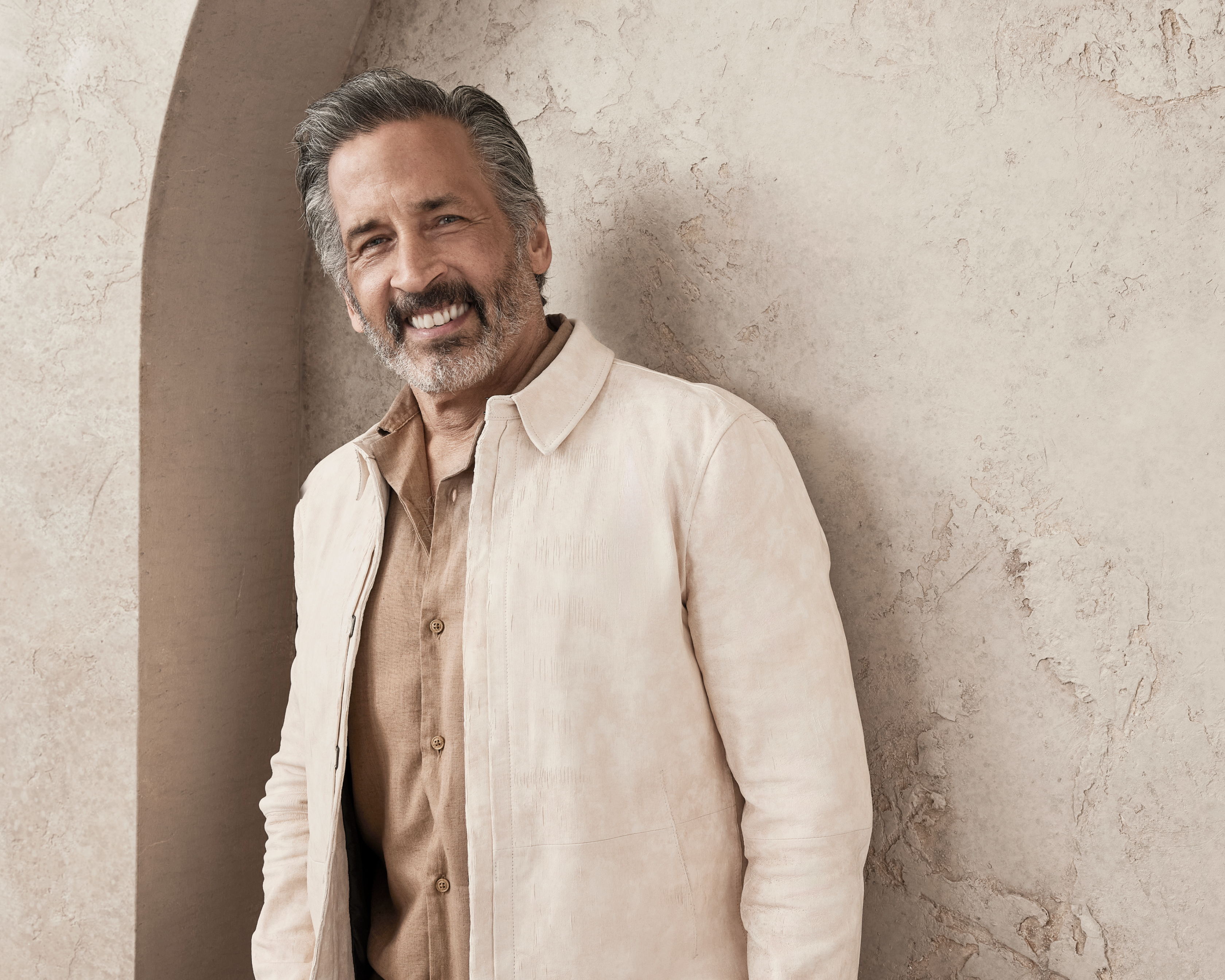 An older man with gray hair and a beard, wearing a casual button-up shirt and jacket, smiling while leaning against a textured wall. Names unknown