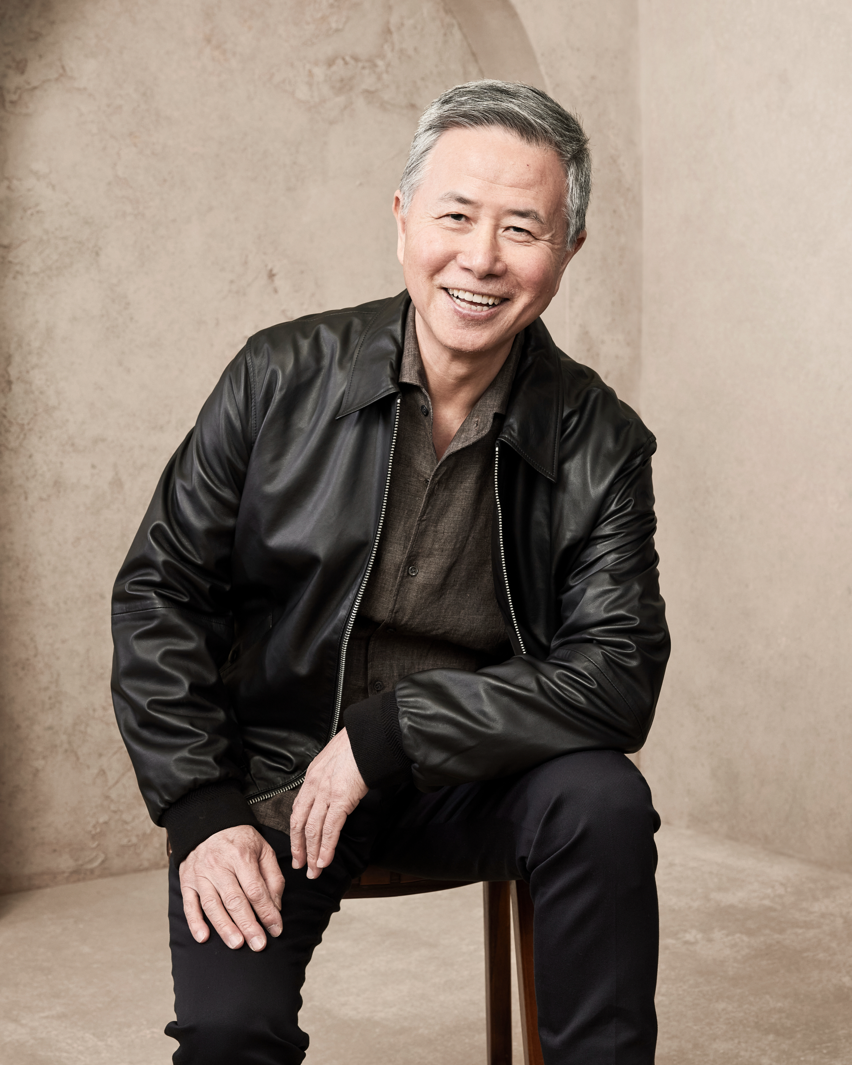 A man is smiling and sitting on a stool. He is wearing a black leather jacket over a dark shirt and dark pants