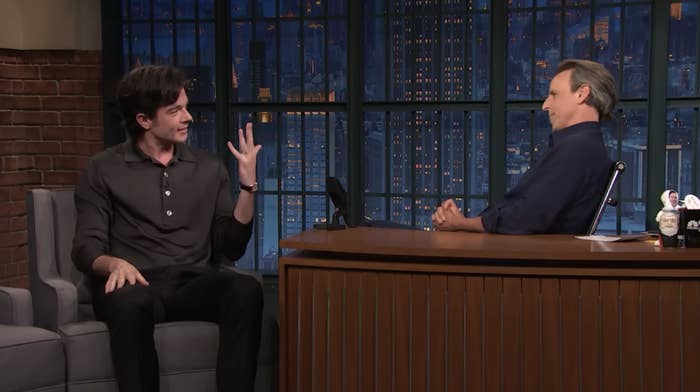 Seth Meyers interviewing Penn John Mulaney on a talk show set, both seated with Penn gesturing animatedly