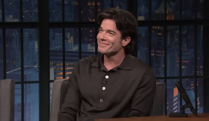 John Mulaney smiles while seated on set during a talk show interview. He wears a dark button-down shirt
