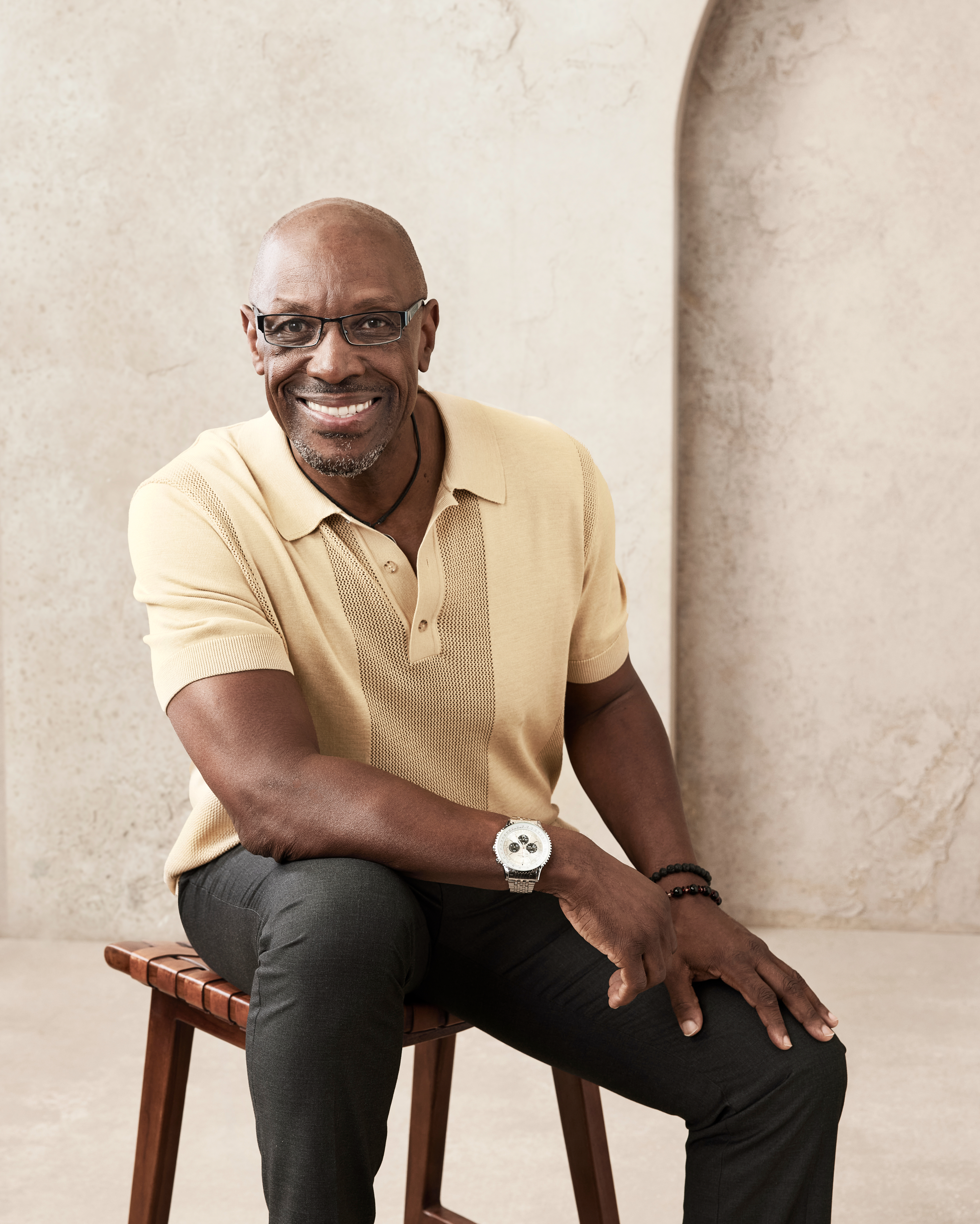 Isaac C. Singleton Jr. is sitting on a wooden stool, wearing a short-sleeve polo shirt and dark pants, smiling at the camera