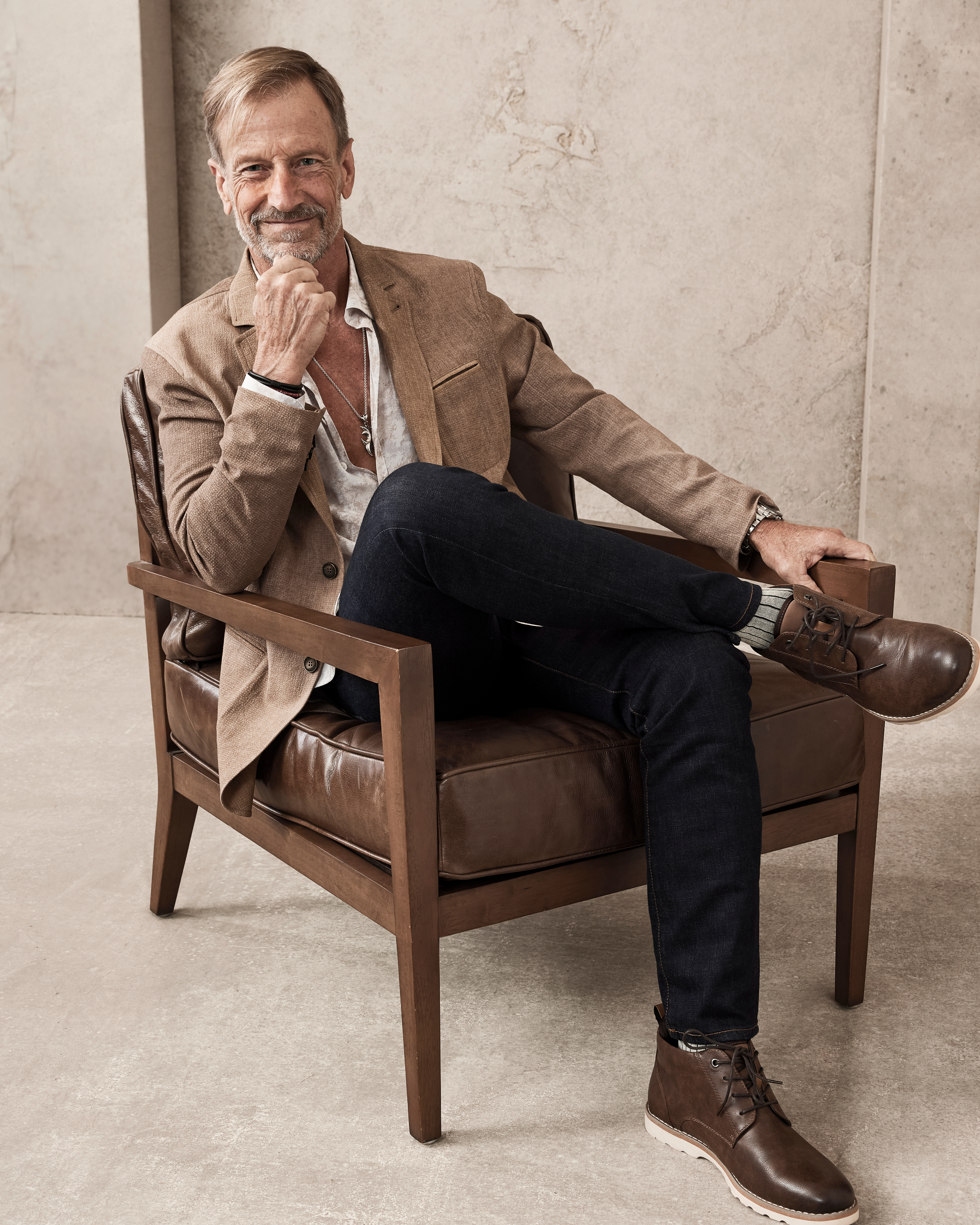 A man poses confidently while seated in a leather chair, wearing a beige blazer, a light shirt, dark jeans, and brown boots. I don't know his name