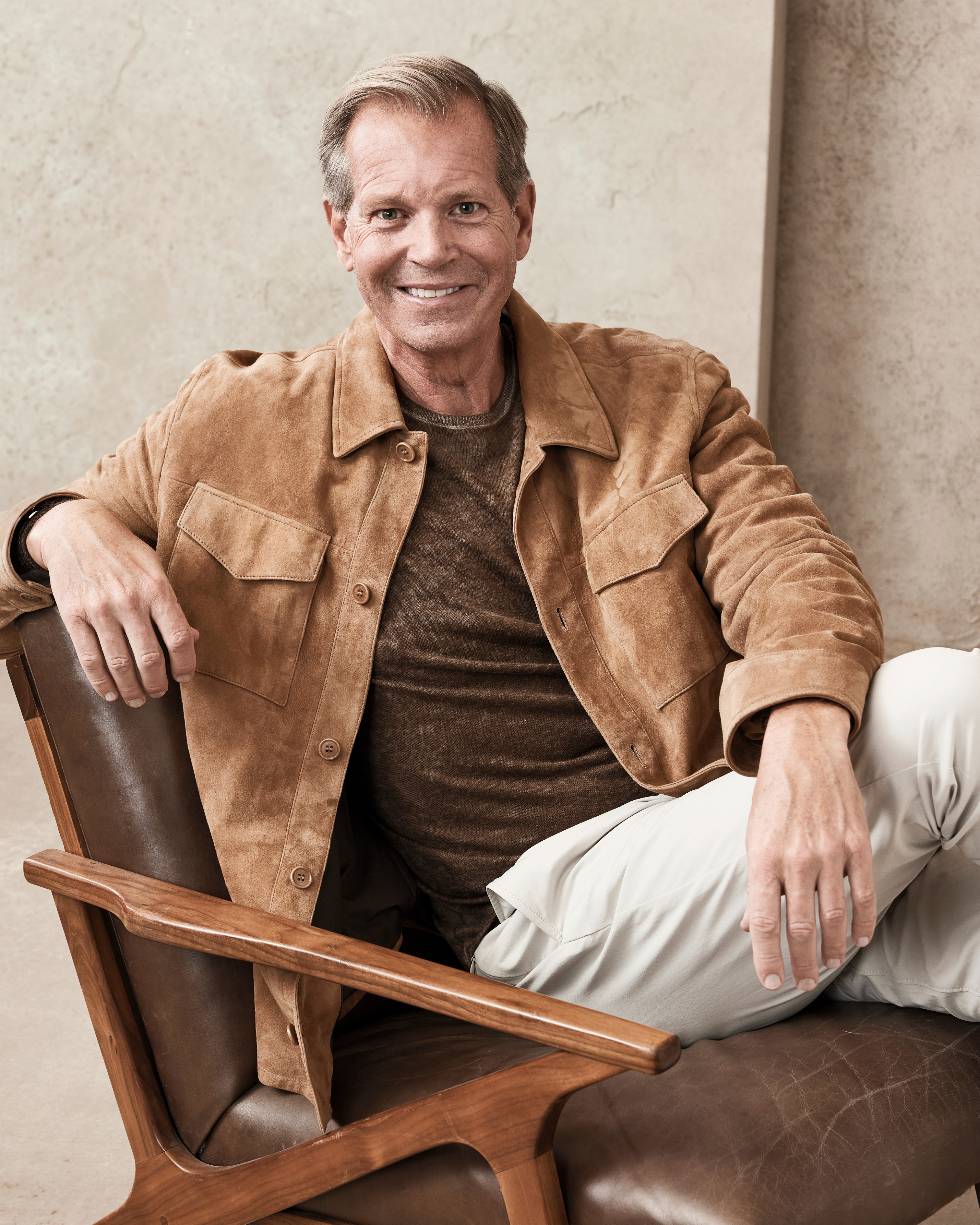 A man sits comfortably on a chair, smiling at the camera. He wears a casual brown jacket over a brown shirt and light-colored pants