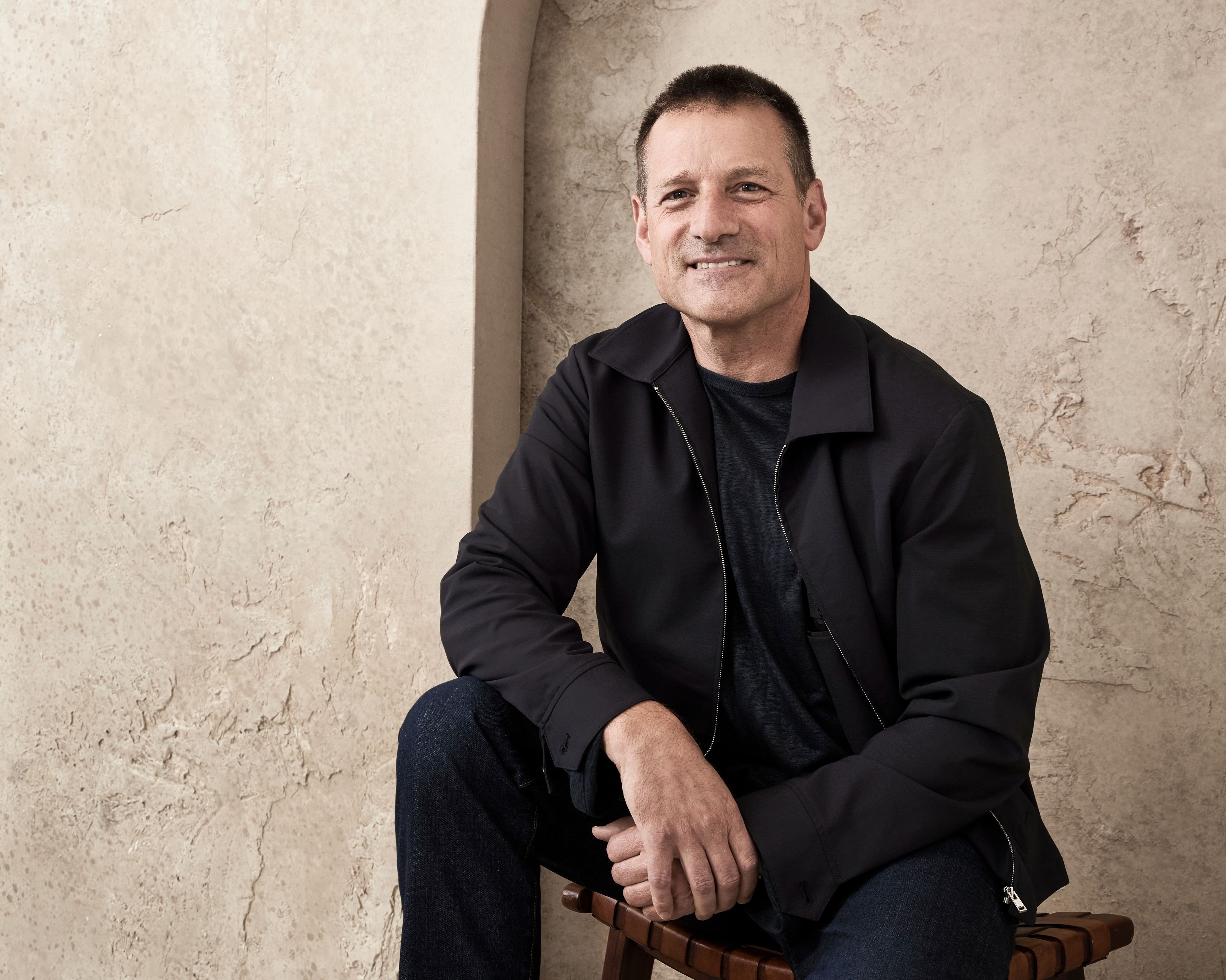 Middle-aged man smiling, wearing a dark jacket and black shirt, seated on a wooden chair against a textured beige wall