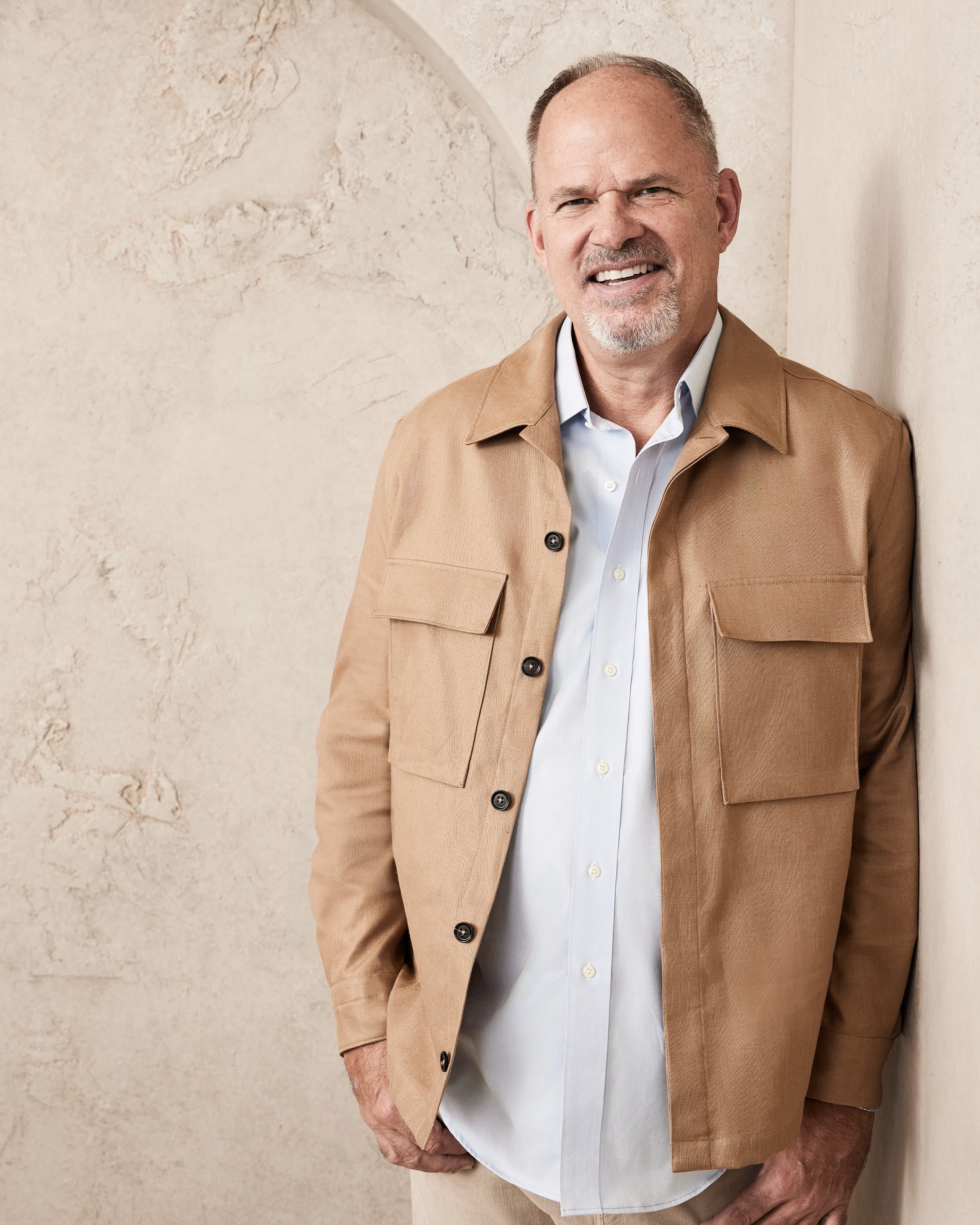 A man with short hair, mustache, and goatee smiles while leaning against a textured wall. He is wearing a light-colored button-up shirt and a tan jacket