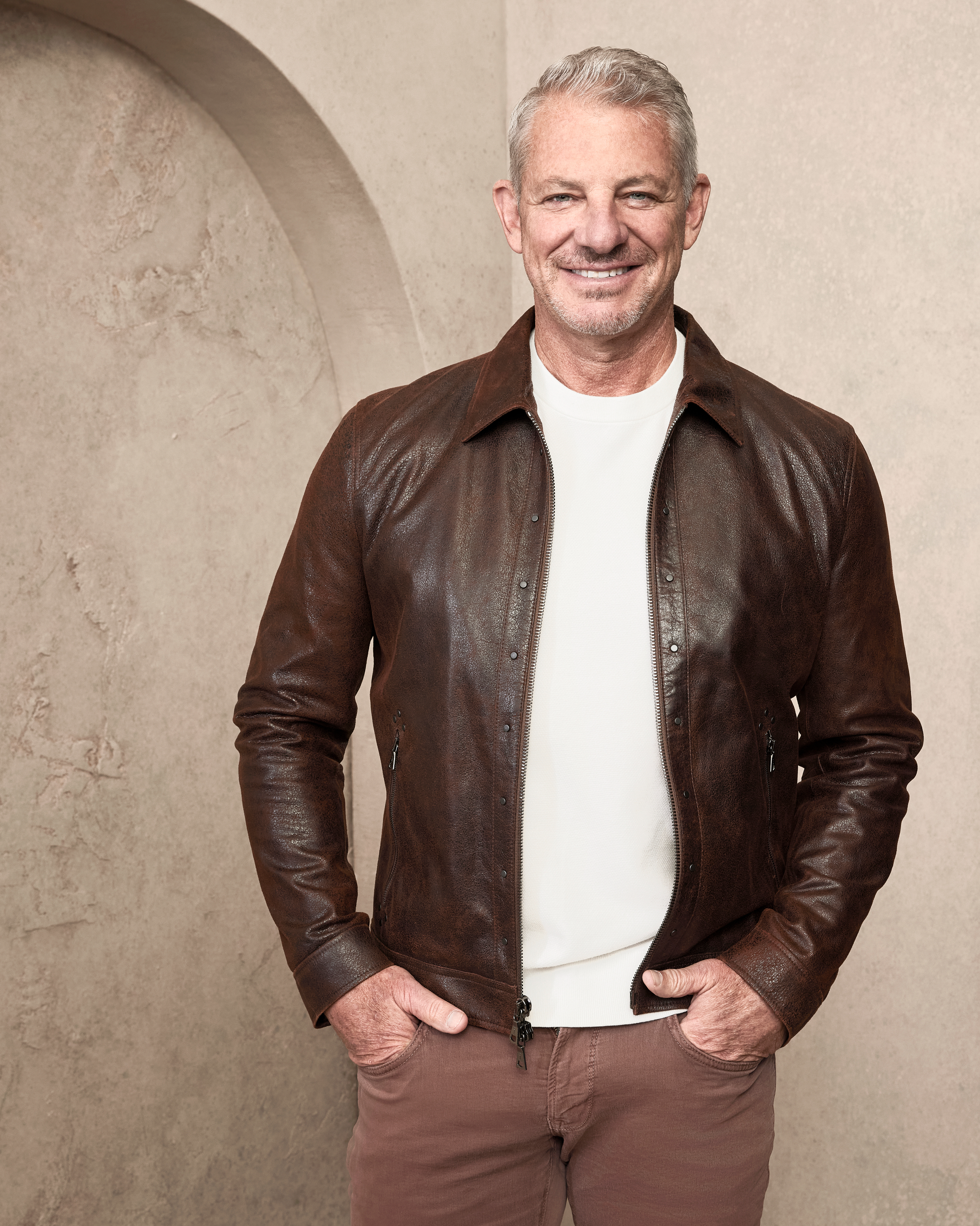 Man in casual attire with a white shirt and brown leather jacket smiling against a neutral background. Name unknown