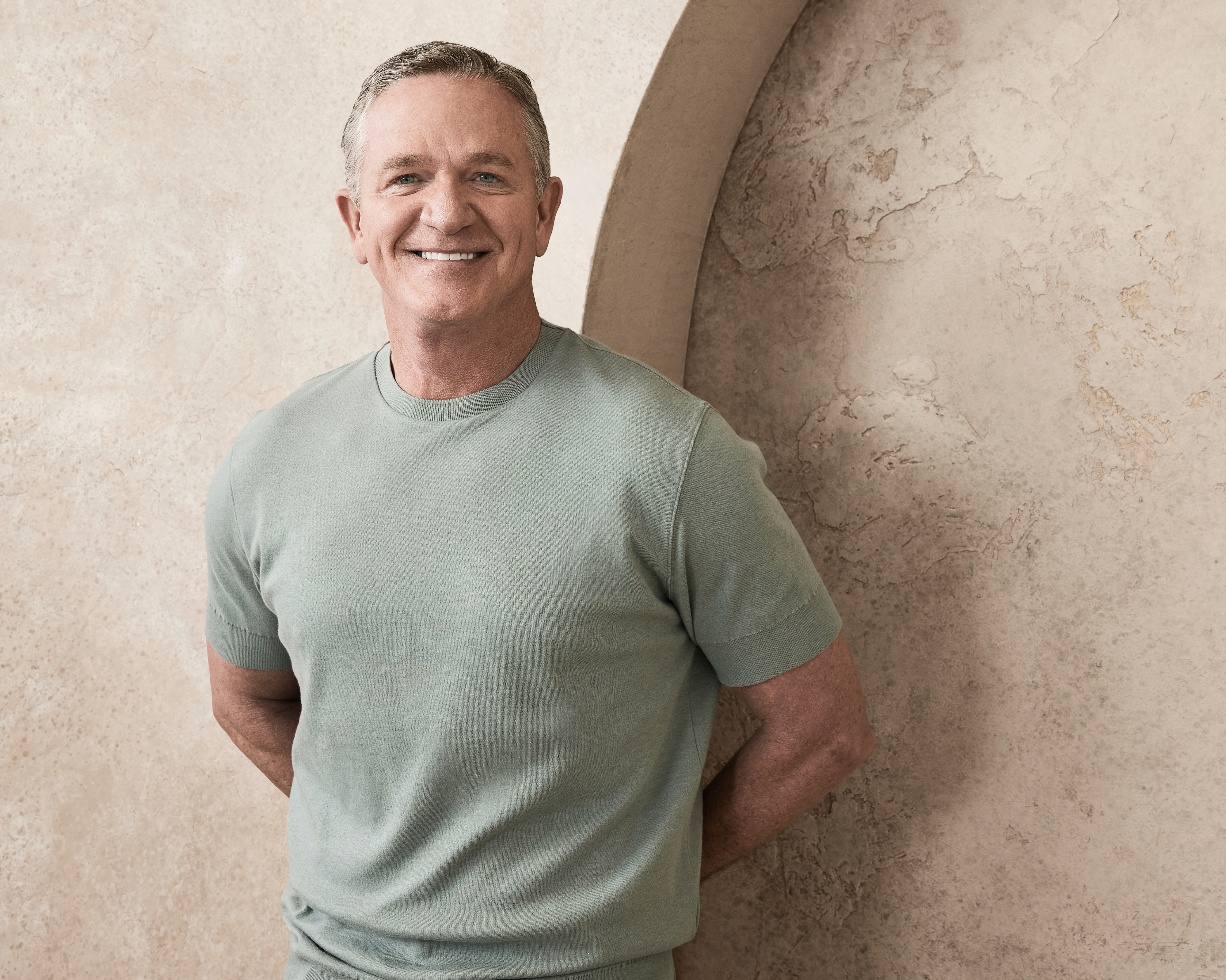 A man standing against a textured wall, smiling, wearing a short-sleeve shirt