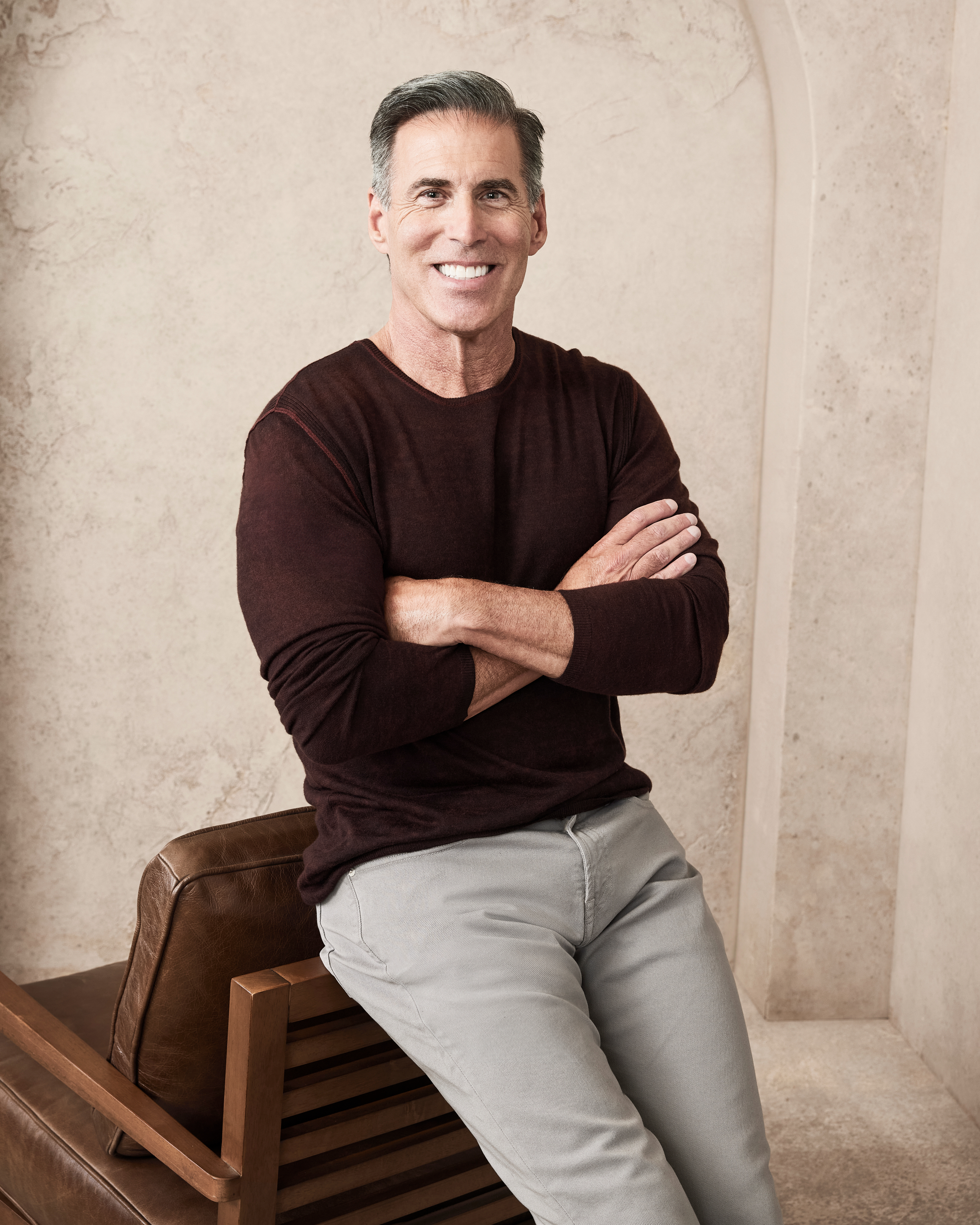 A man with short gray hair, wearing a long-sleeved shirt and light pants, sits on a chair with arms crossed and smiling. Names are not provided