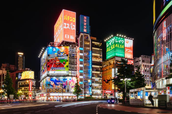 Night view of a vibrant Tokyo street with bright signs and billboards. Crowded buildings showcase various ads, including anime characters and store names