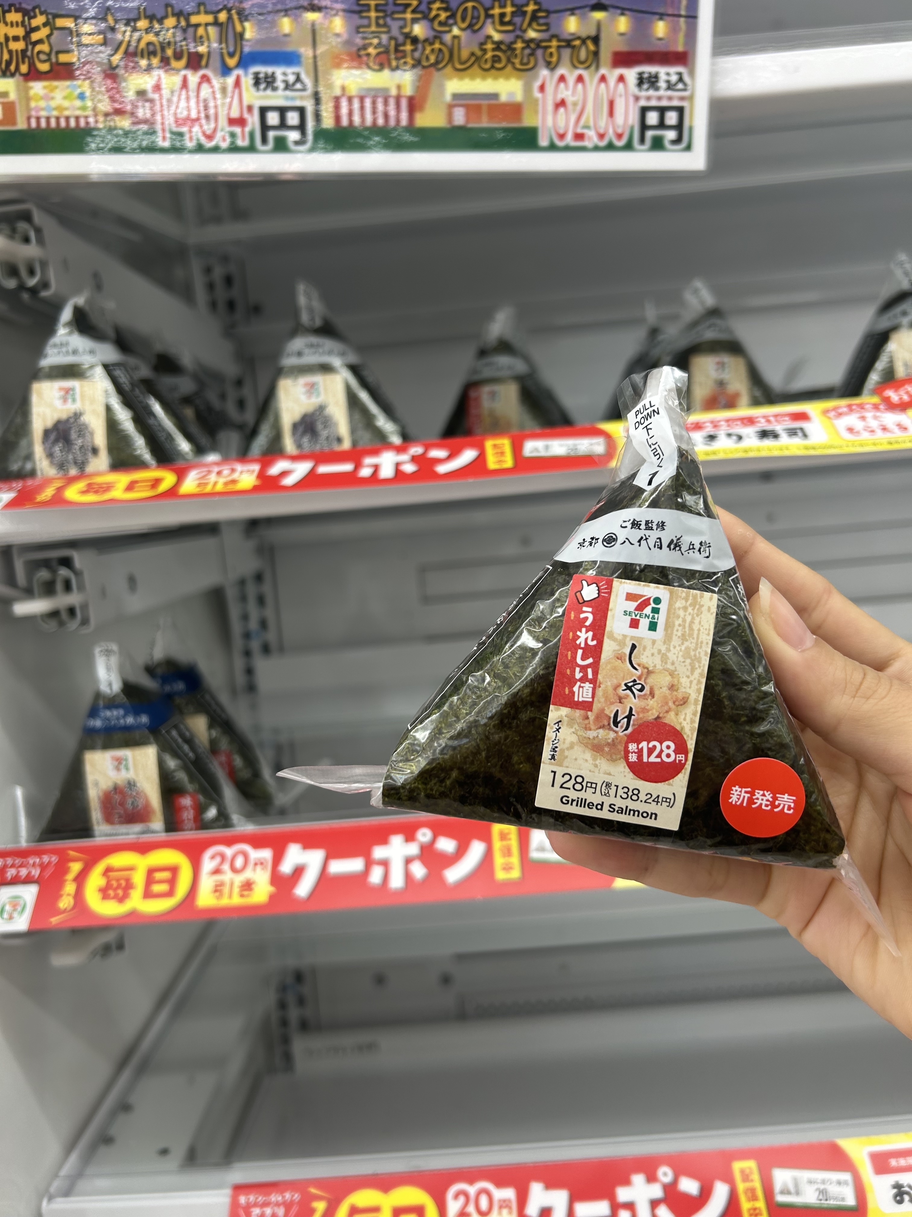 A hand holding a grilled salmon onigiri (Japanese rice ball) in a convenience store. Various other onigiris are displayed on the shelf with price tags