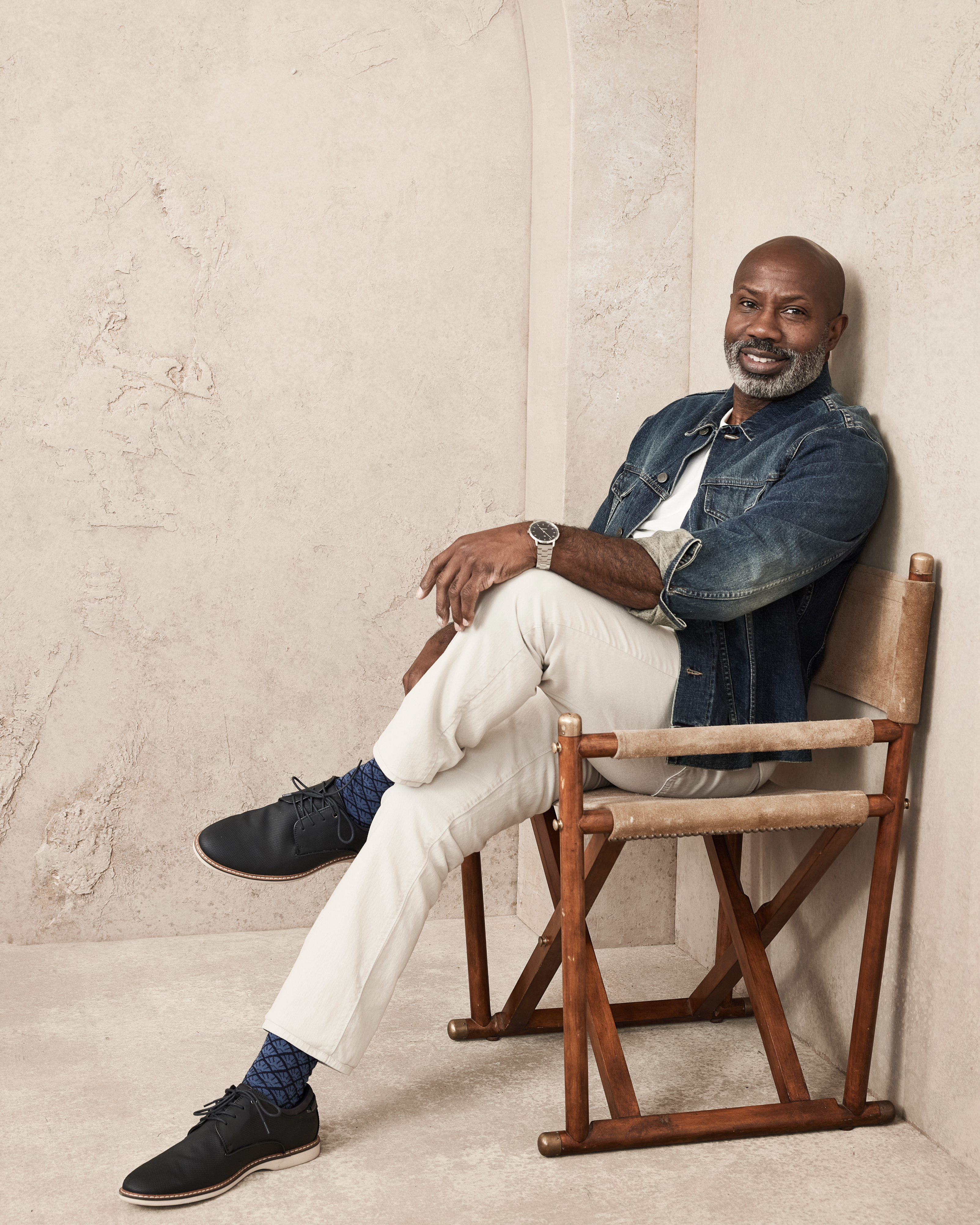 A man wearing a denim jacket, white pants, and black shoes sits on a wooden chair, smiling against a neutral background