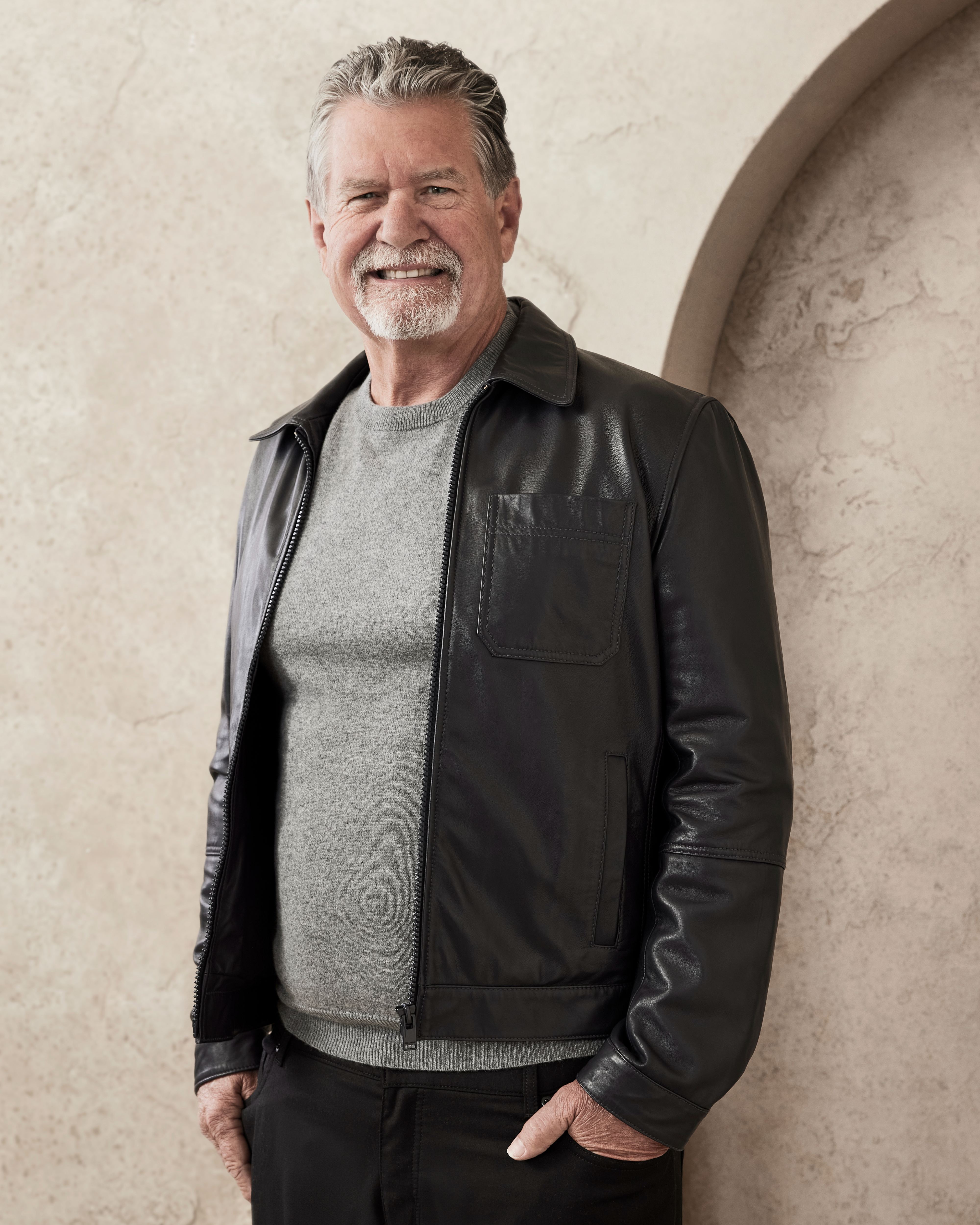 A man, smiling, is wearing a leather jacket over a sweater, standing against a neutral, textured background