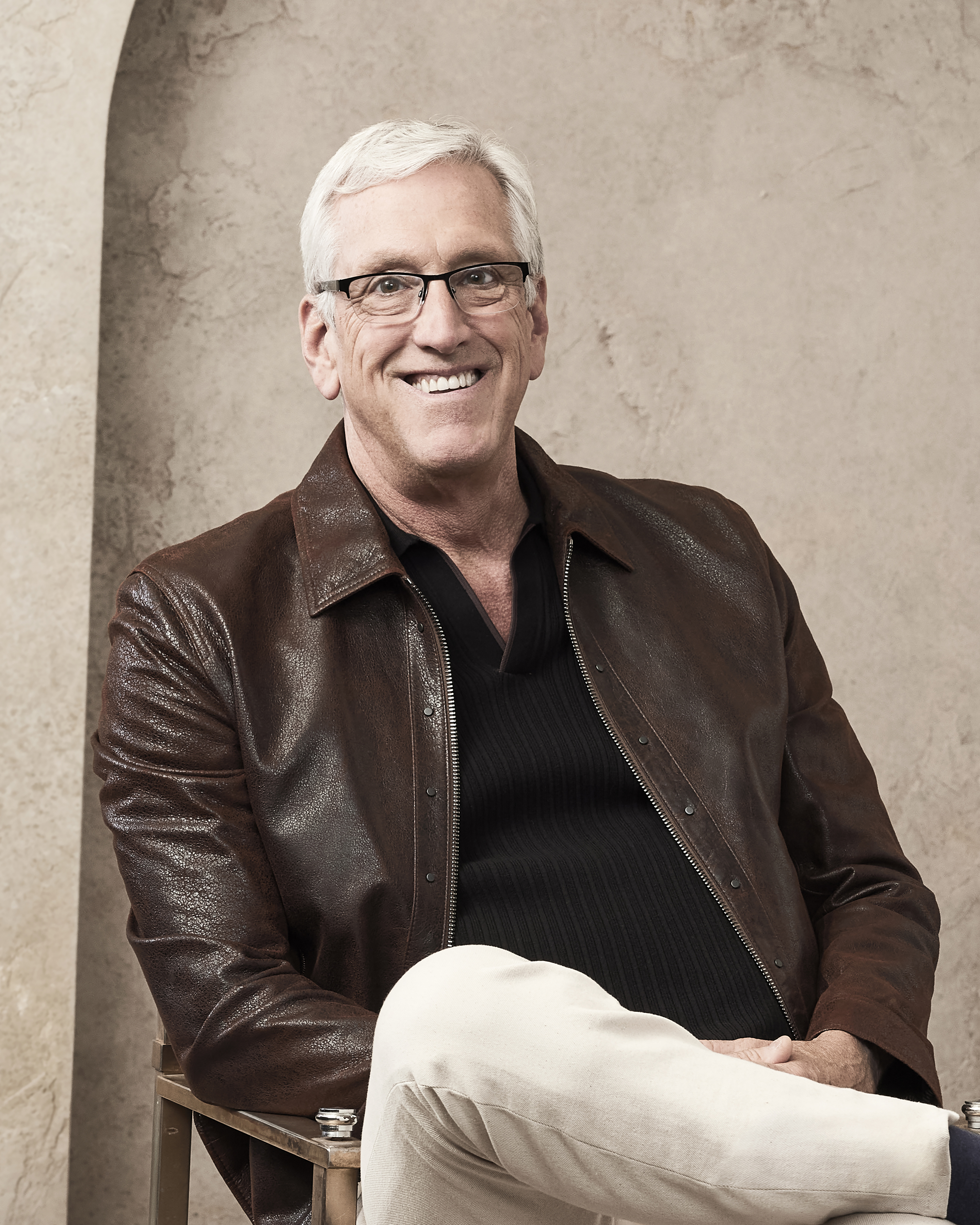 A man with short grey hair and glasses, wearing a brown leather jacket over a black shirt, poses while sitting on a chair with his legs crossed