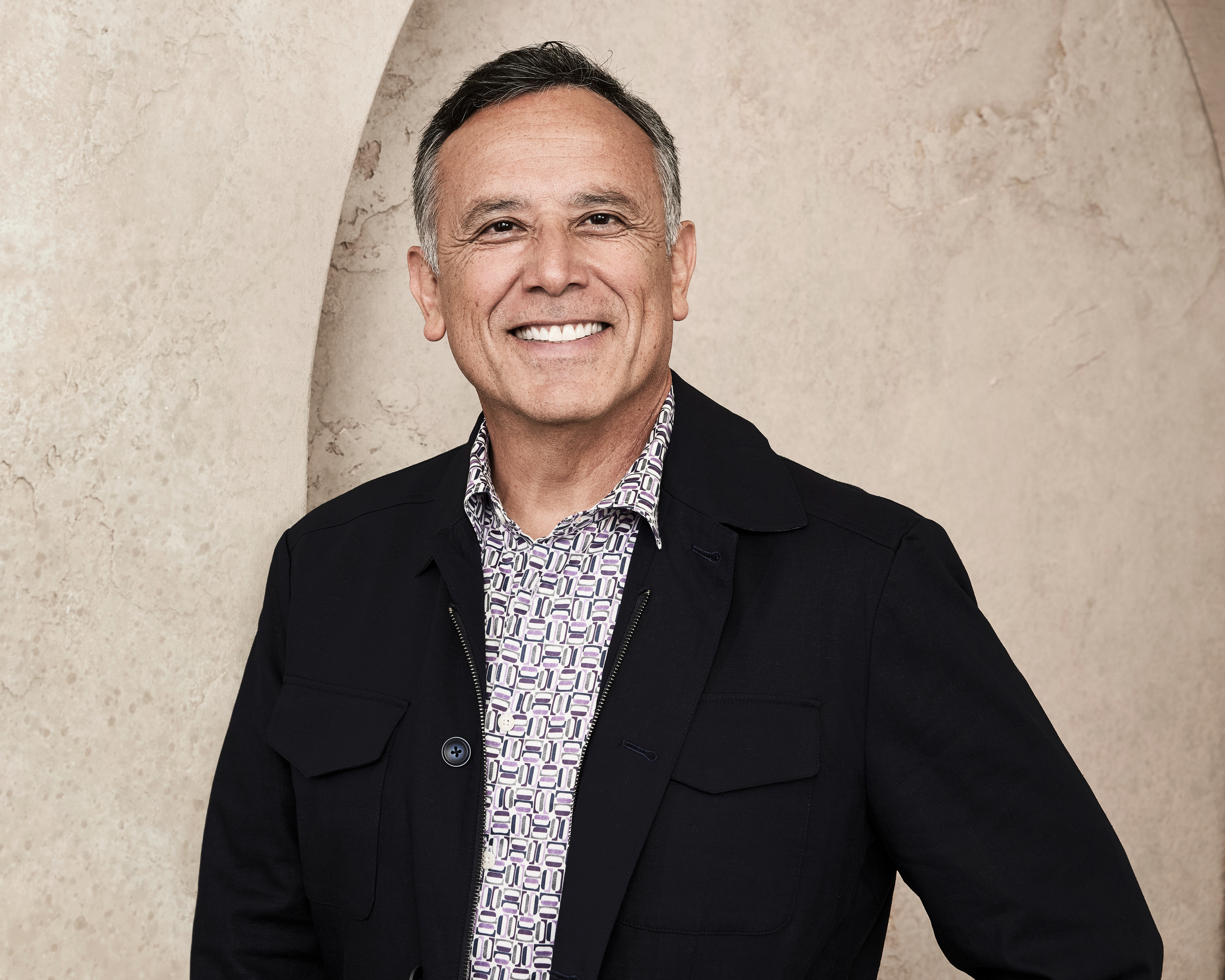 An individual smiling in front of a neutral background, wearing a patterned shirt under a dark jacket