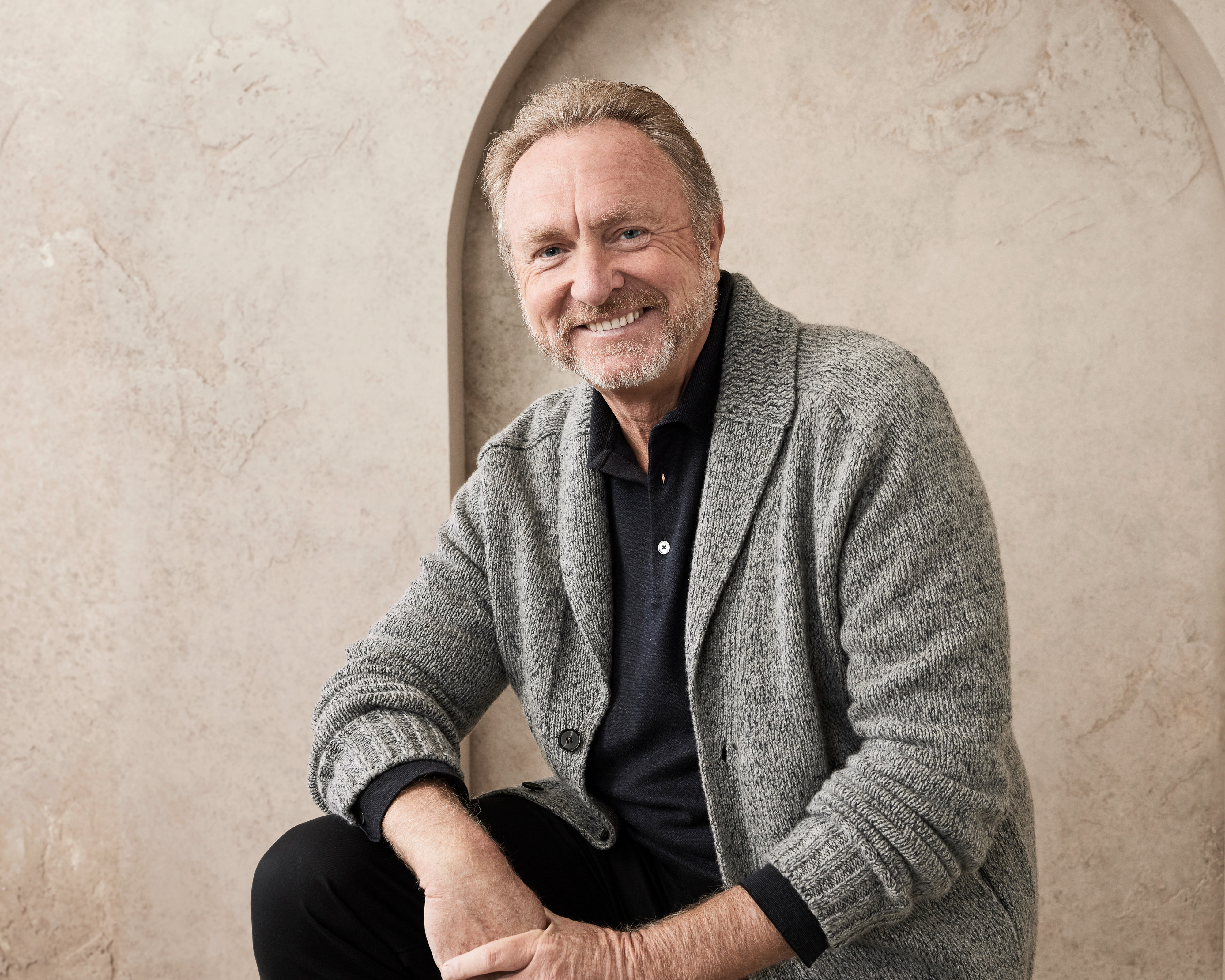 Man sits and smiles, wearing a casual open cardigan over a collared shirt in front of a textured wall background. Unknown person