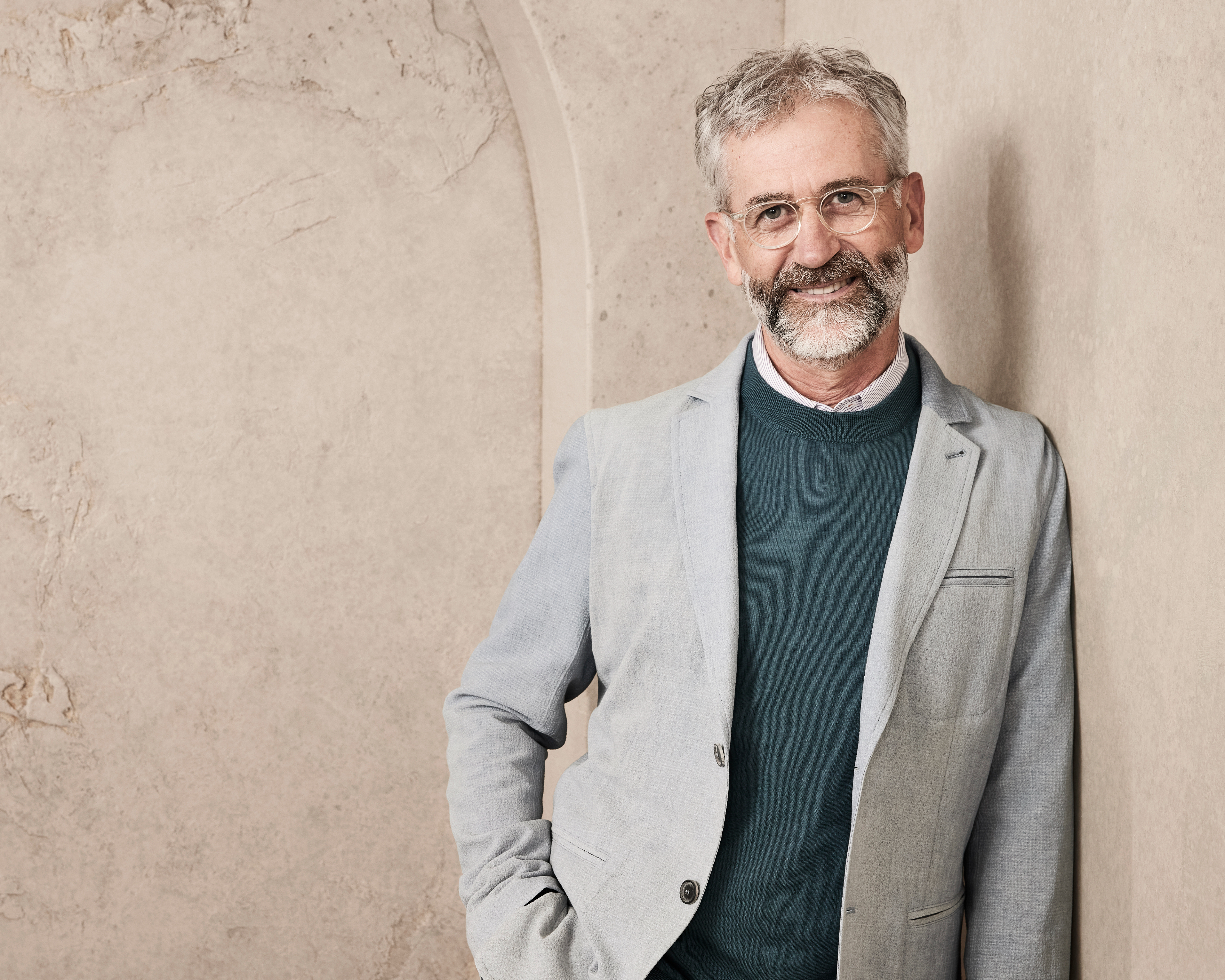 A man with glasses and a short beard stands against a textured wall, smiling. He is wearing a light blazer over a sweater