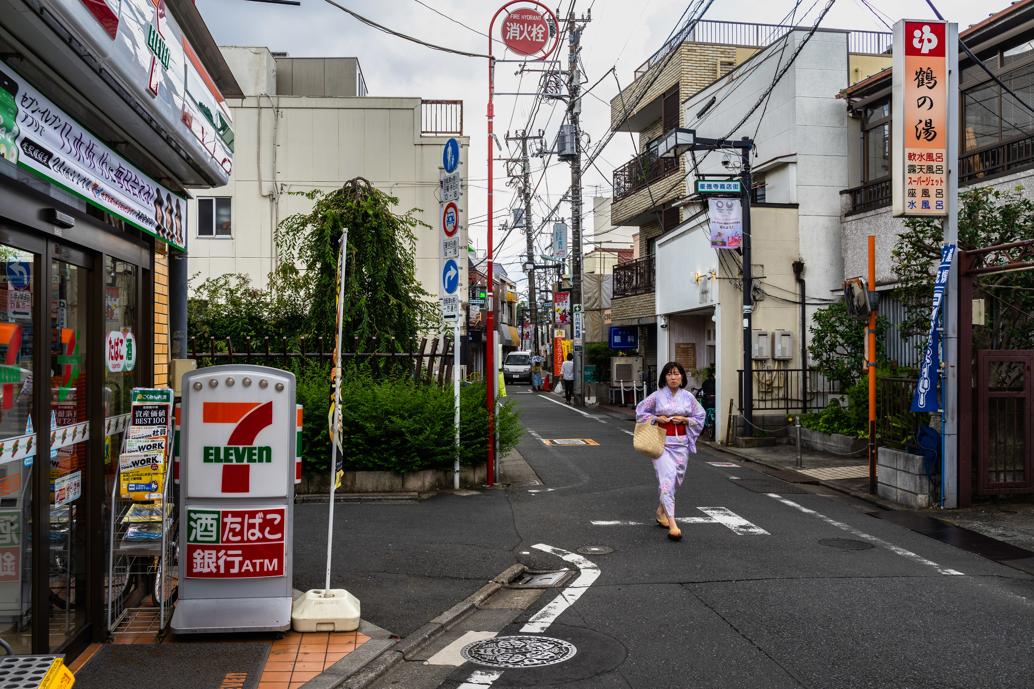 A person in traditional Japanese attire walks down a narrow street lined with various shops and signs, including a 7-Eleven
