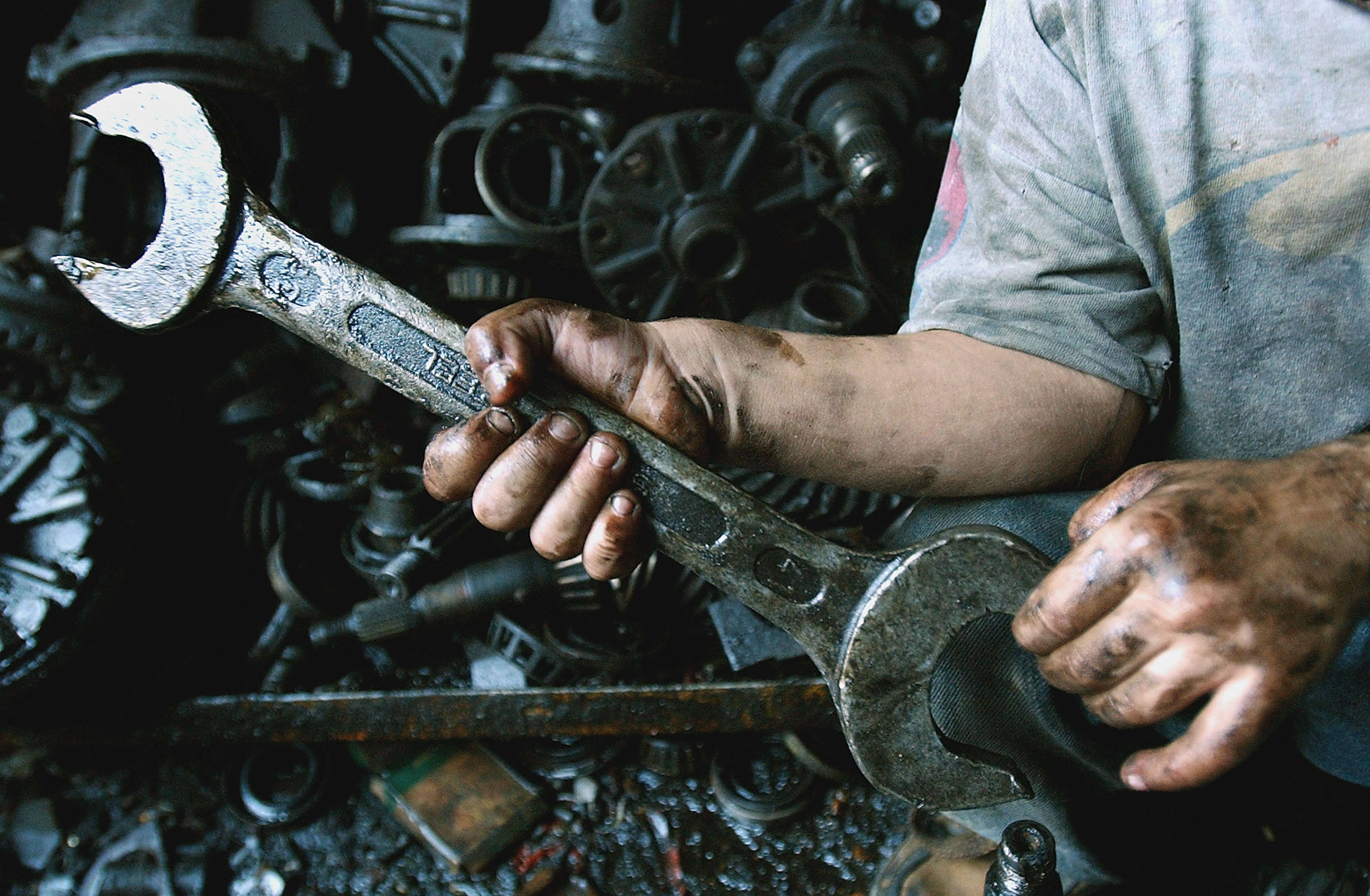 A person holding a large wrench with dirty, greasy hands, surrounded by various mechanical parts, indicating manual labor or mechanical work