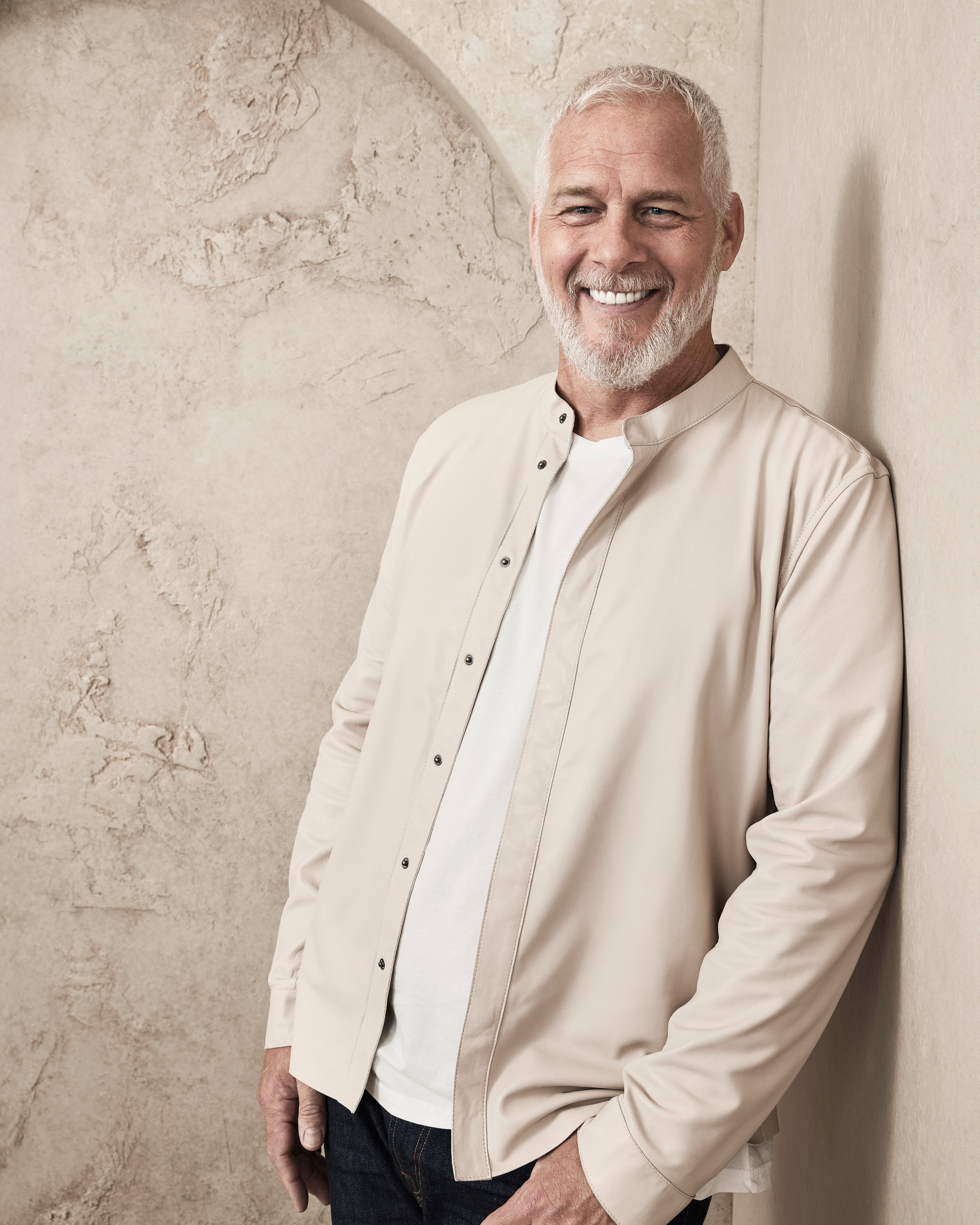 A man with white hair and beard smiles while leaning against a textured wall. He is wearing a white shirt and a light jacket