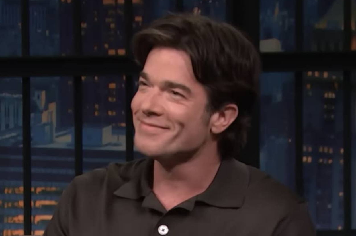 John Mulaney smiling during an interview on a nighttime talk show set with city skyline in the background