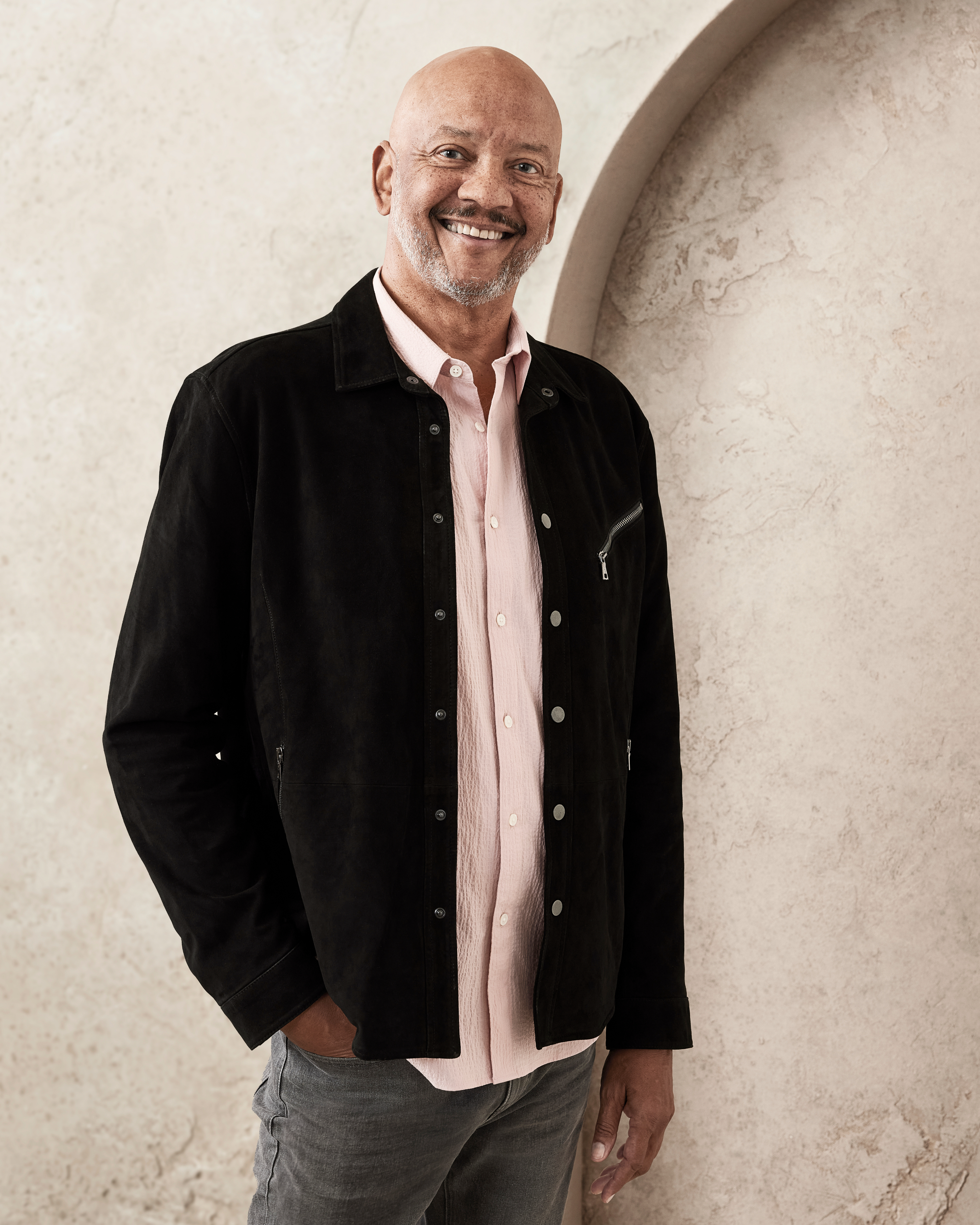 Man smiling, wearing a casual outfit with a black jacket, light shirt, and gray pants, standing against a textured wall. I don't know who they are