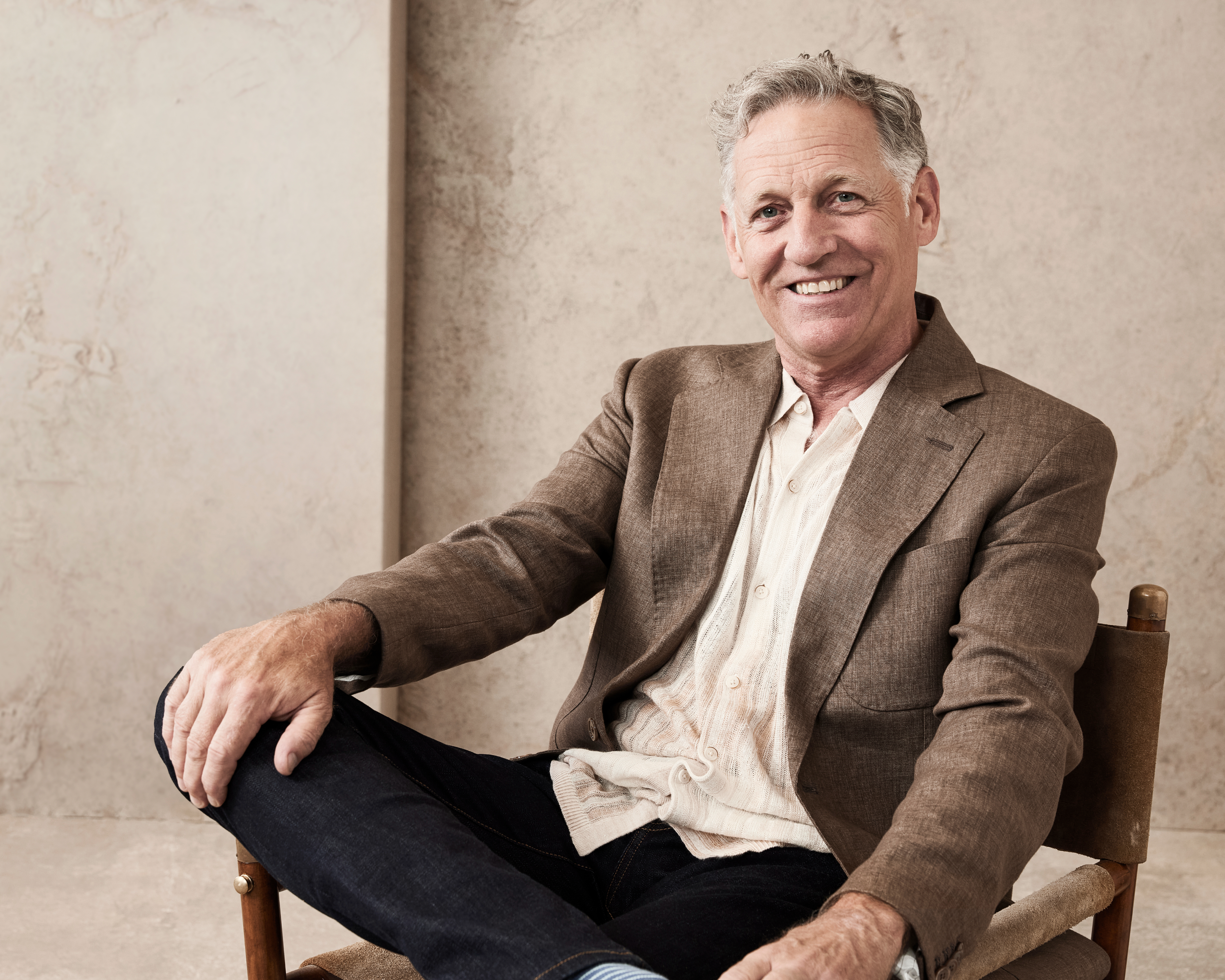 Older man with short gray hair, wearing a light shirt, blazer, and jeans, sits casually on a chair, smiling at the camera