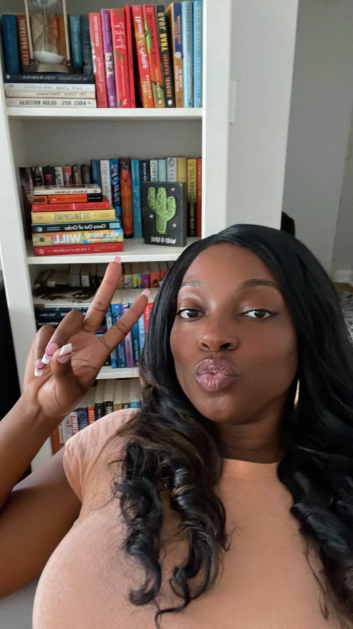 A person poses making a peace sign in front of a bookshelf filled with various books