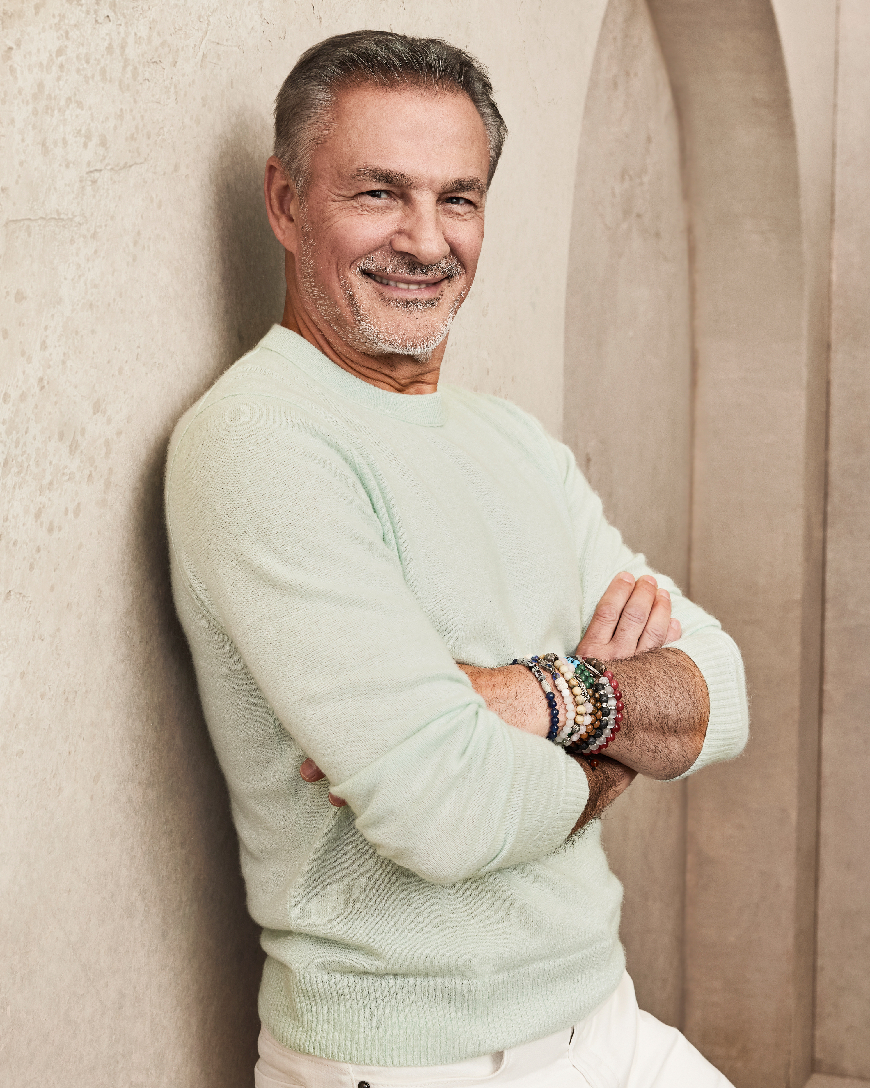 Man leaning against a wall, smiling with arms crossed. He is wearing a light sweater and beaded bracelets