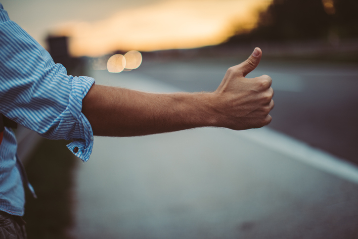 Person's arm giving a thumbs-up while standing by the side of a road at sunset