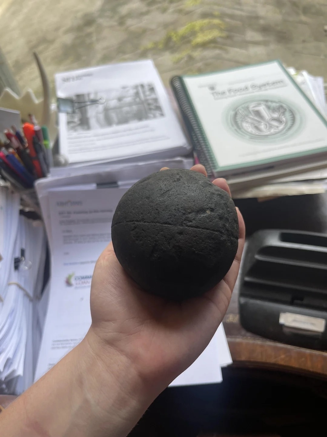 A hand holds a dark, round object over a cluttered desk with papers, books, and a pen holder in the background