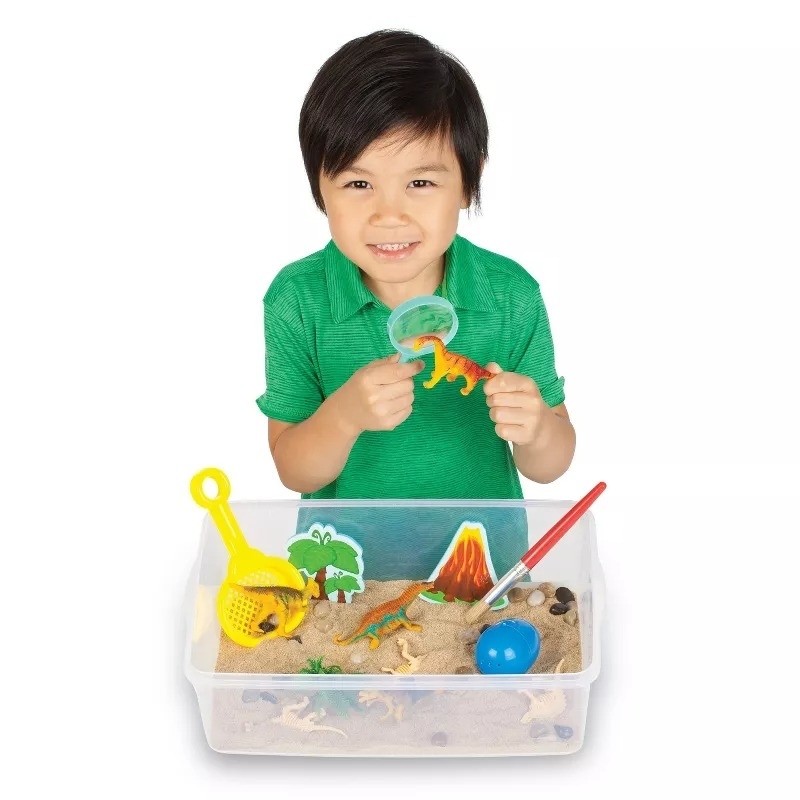 Child playing with a dinosaur-themed sensory bin filled with sand, toys, and tools