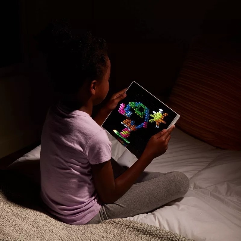 Young child in pajamas holding a Lite-Brite creation while sitting on a bed