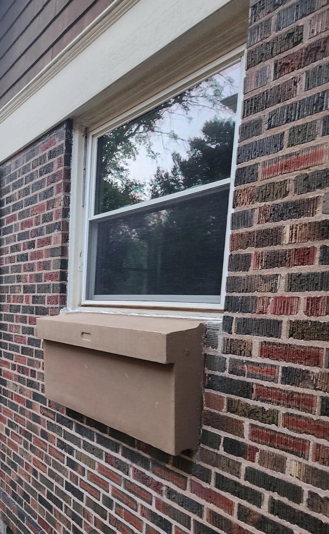 A red-brick wall with a window and an overhanging brown box covers the window's lower portion. Trees are visible in a reflection in the window