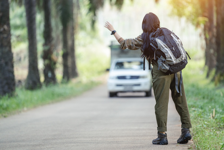 A person with long hair and a large backpack is hitchhiking on a rural road, waving at an approaching white vehicle