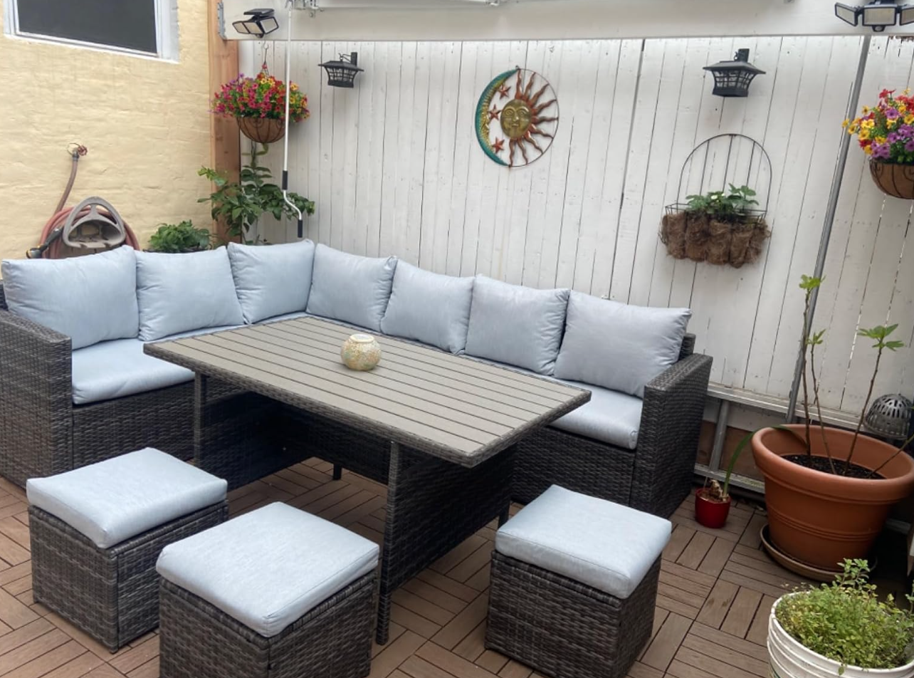 Patio set with a wicker bench, light cushions, a rectangular table, and matching stools in a cozy garden space with hanging plants and wall decor