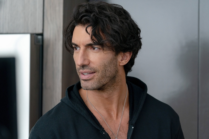 A man with wavy dark hair and a short beard wearing a dark hoodie and layered necklaces, looking off to the side