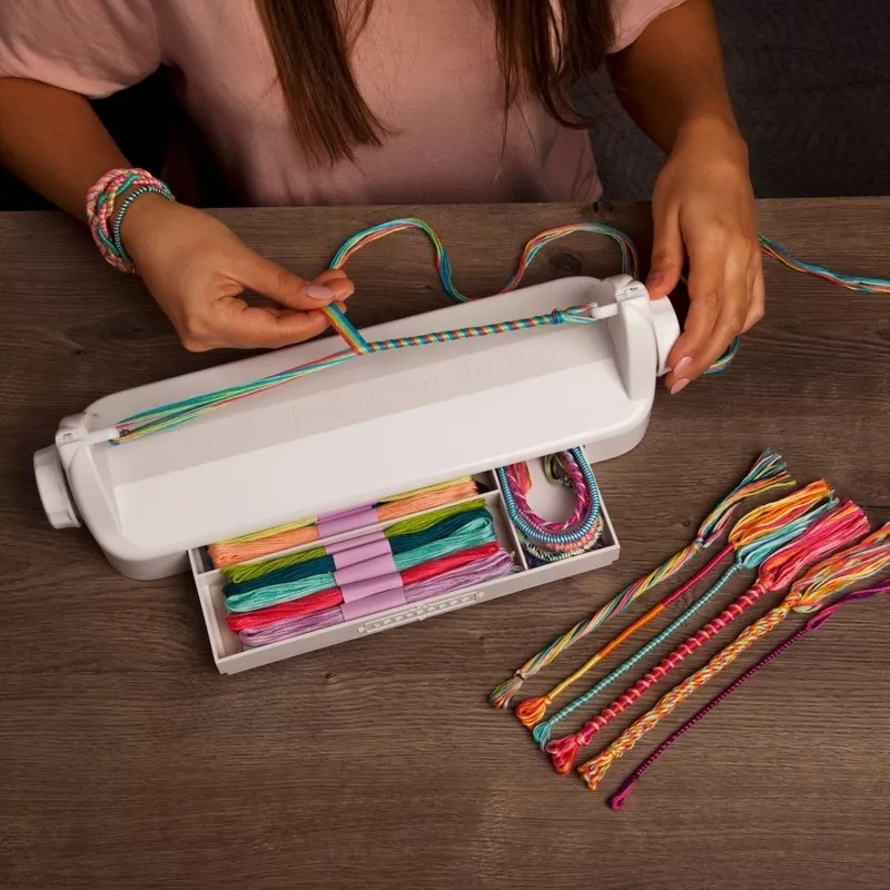 A person making braided friendship bracelets using a DIY bracelet maker, with multicolored thread kits laid out on a wooden table