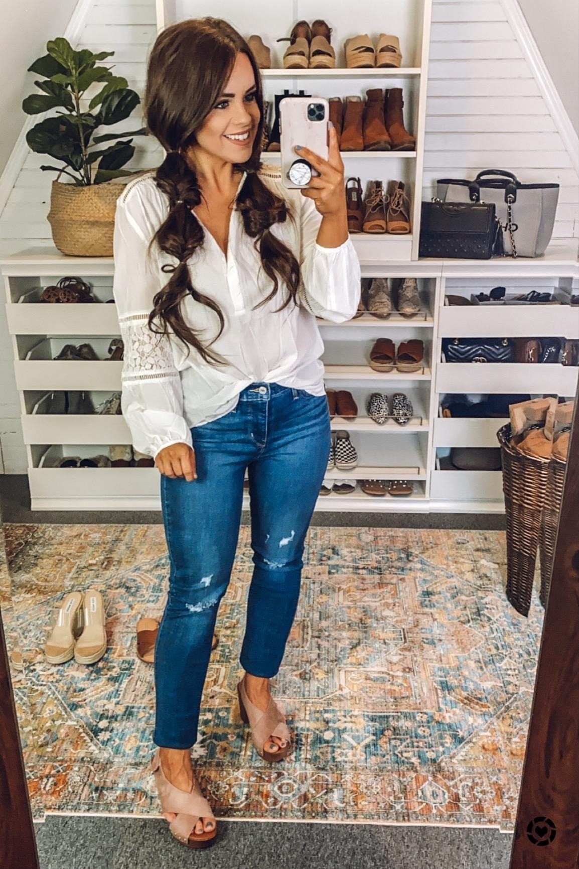 a reviewer in a white blouse and jeans takes a mirror selfie in a walk-in closet filled with shelves of shoes and handbags