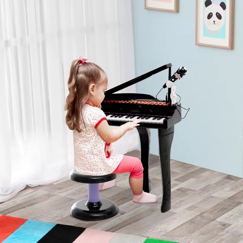 A young child sits at a small piano, playing music in a room with framed pictures on the wall