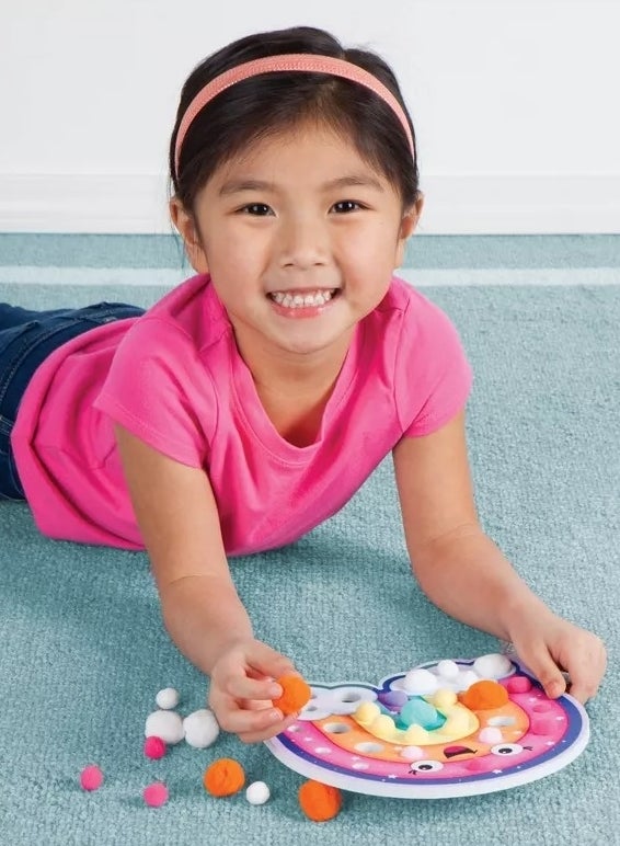 A child smiles while lying on a carpet and playing with a colorful pom-pom sorting toy