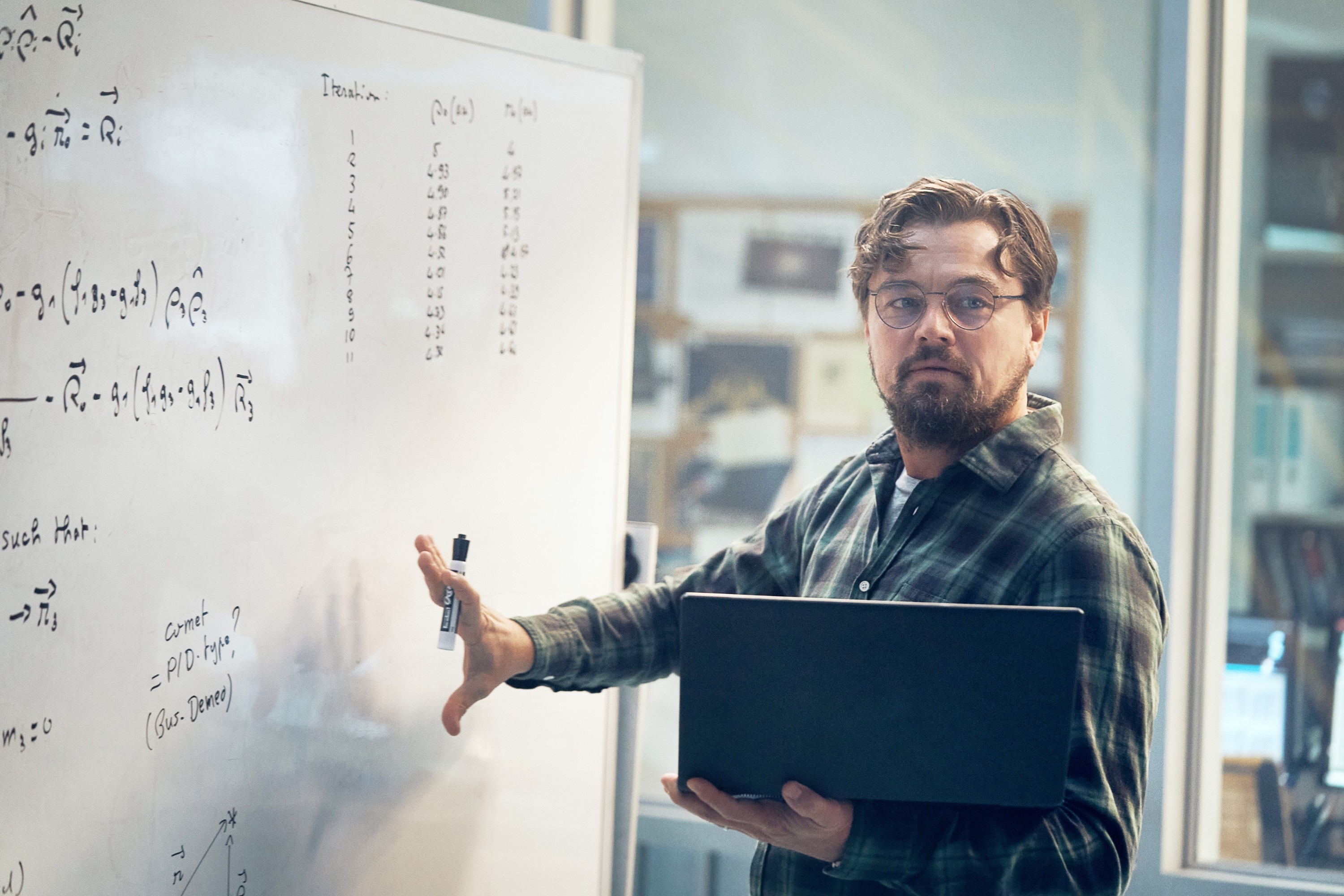 Leonardo DiCaprio, wearing glasses and a plaid shirt, stands by a whiteboard with mathematical equations, holding a laptop, and pointing to the board in &quot;Don't Look Up&quot;