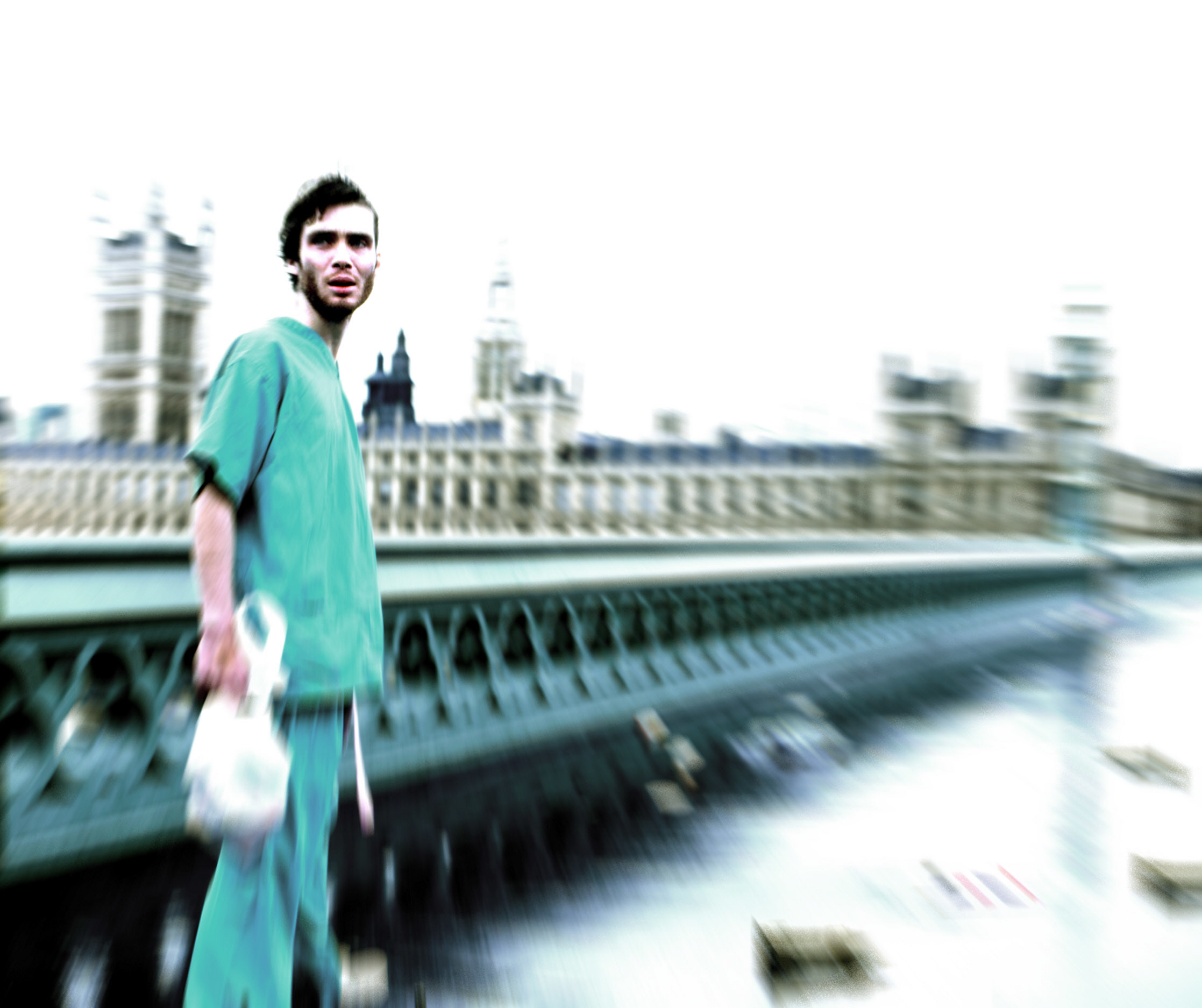 Cillian Murphy in medical scrubs stands on a bridge in front of blurred historic buildings in &quot;28 Days Later&quot;