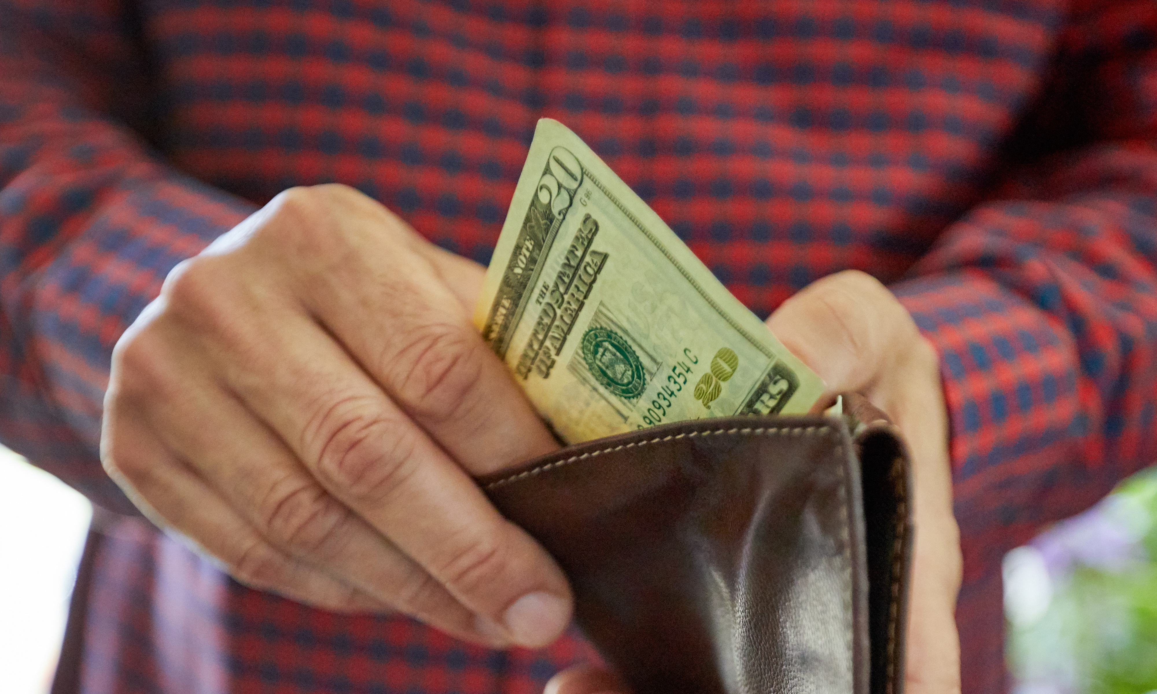 A person in a plaid shirt places a $20 bill into a brown leather wallet